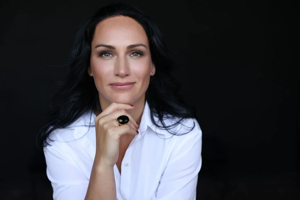 A woman with black hair and fair skin, wearing a white shirt, posing against a black background. She is resting her chin on her hand, which has a large black ring.