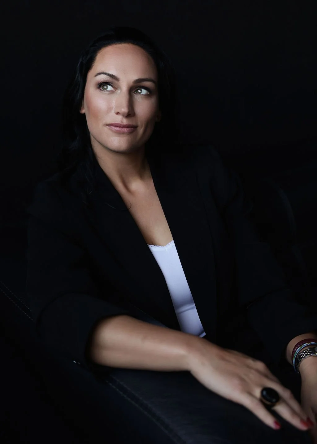 A woman with dark hair and light skin wearing a black blazer and white top, sitting against a black background, looking to the side with a thoughtful expression.