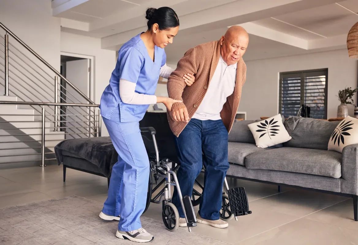 Nurse helping elderly man walk with a wheelchair in a living room.