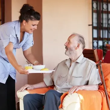A young woman in a blue uniform serving breakfast to an elderly man with a beard in a wheelchair