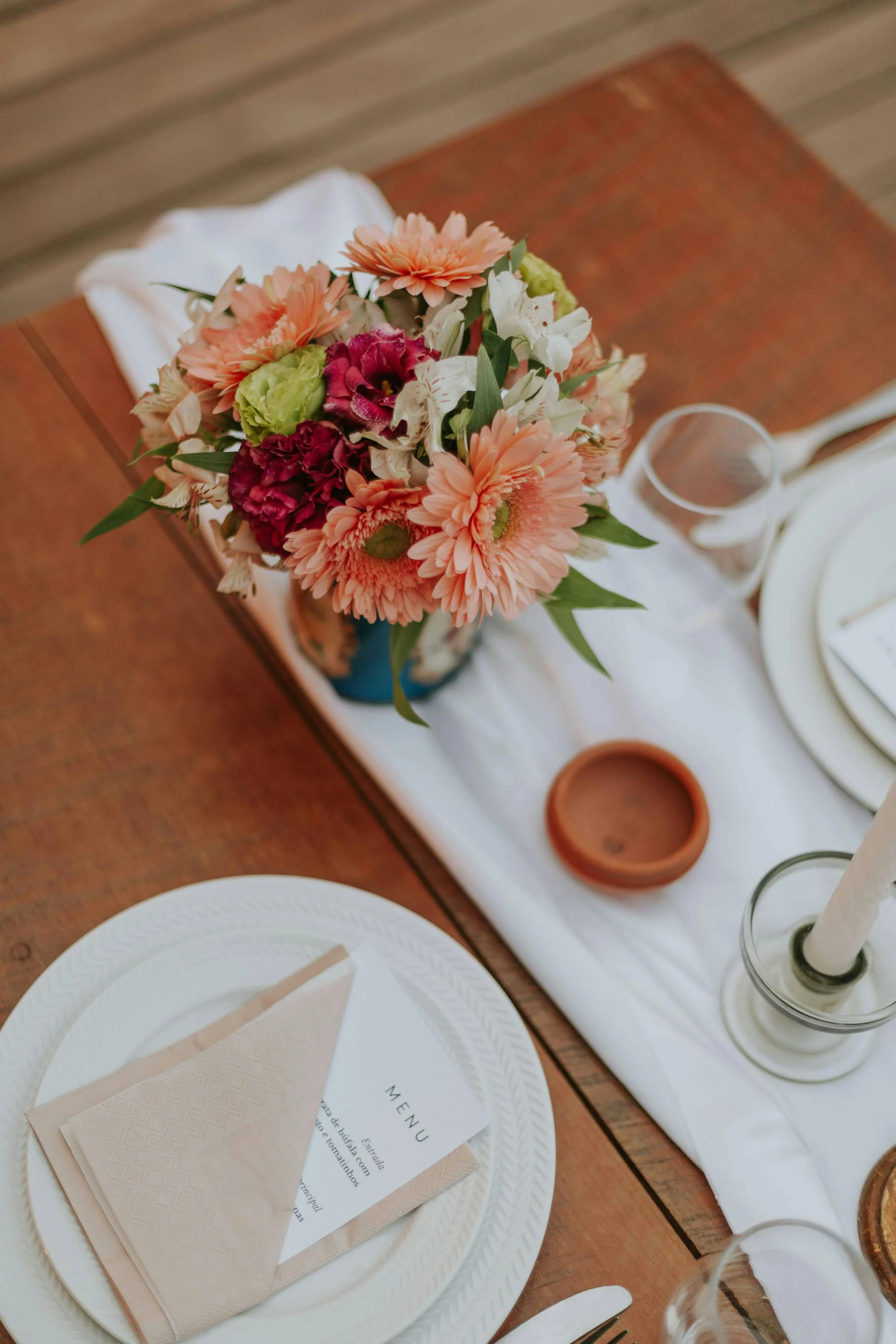 A table setting with a floral centerpiece of pink, white, burgundy, and green flowers, a white plate with a beige napkin and menu, a wine glass, a candle in a holder, and a terracotta dish, all arranged on a white table runner on a wooden table.