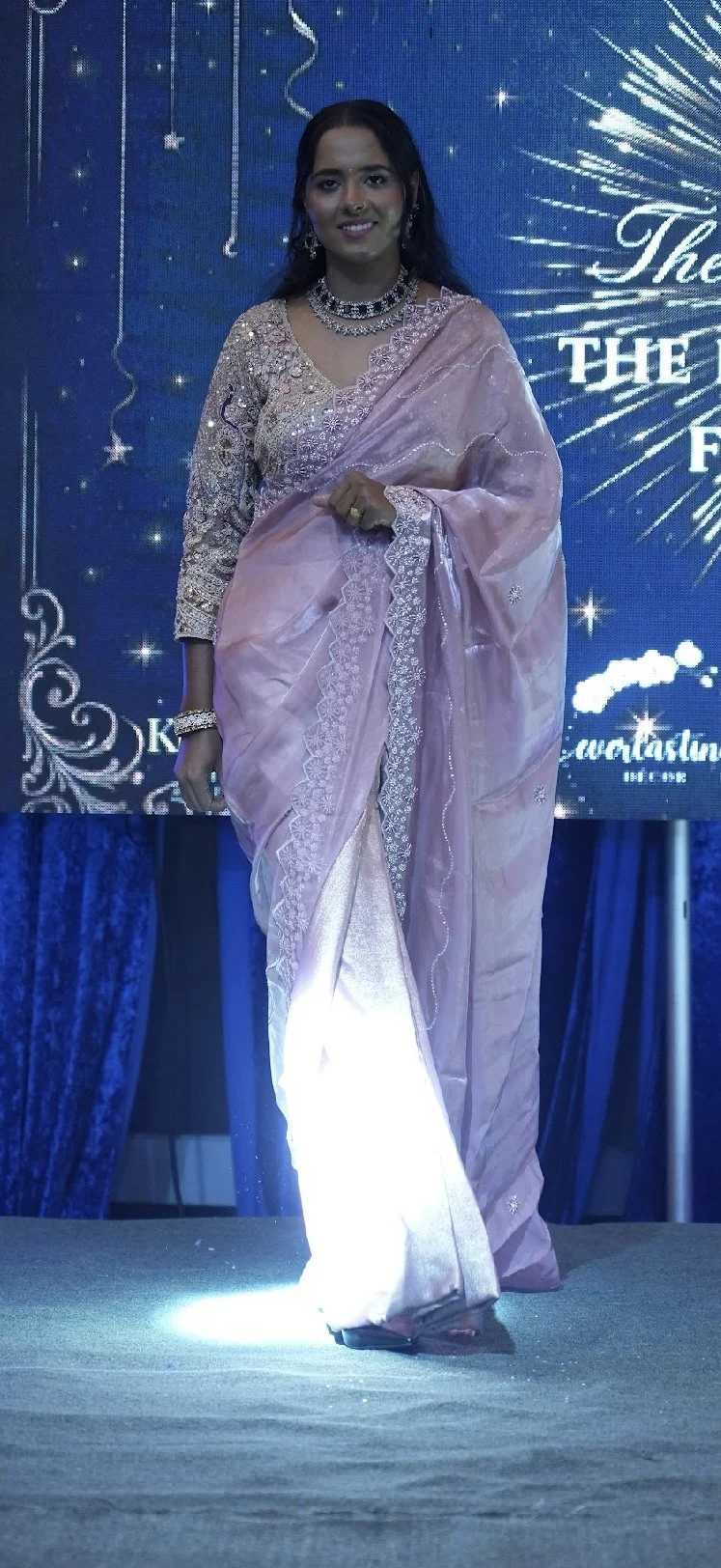A woman wearing a pink traditional Indian saree and jewelry, standing on a stage with a blue background featuring decorative designs and the text 'The' and 'F'.
