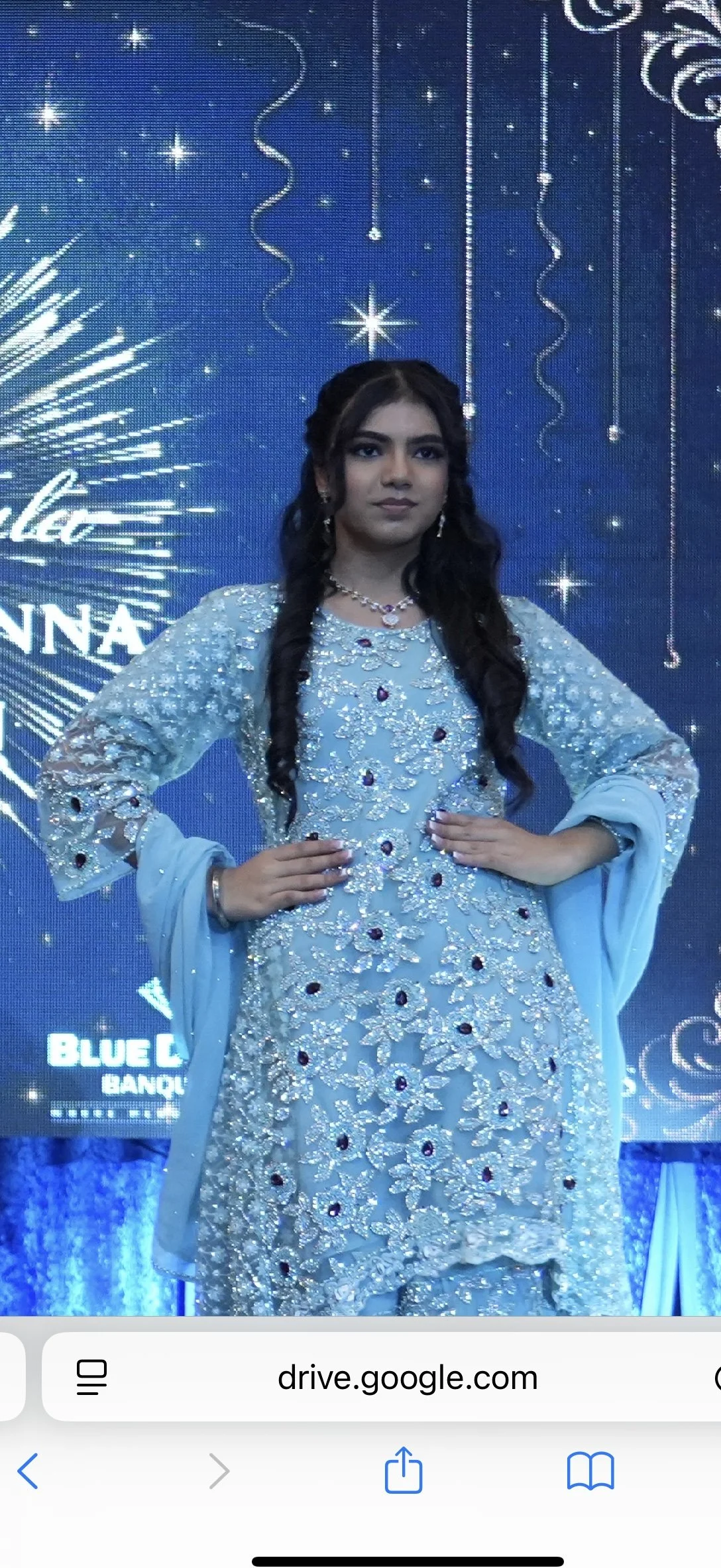 Young woman in an ornate light blue traditional dress with embroidery and gemstones, standing confidently with hands on hips, in front of a festive, starry backdrop.