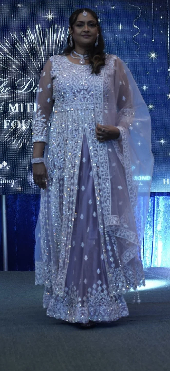 Woman in an elegant, embellished dress with intricate silver detailing at a formal event, standing on a stage with a decorative backdrop featuring stars and fireworks.