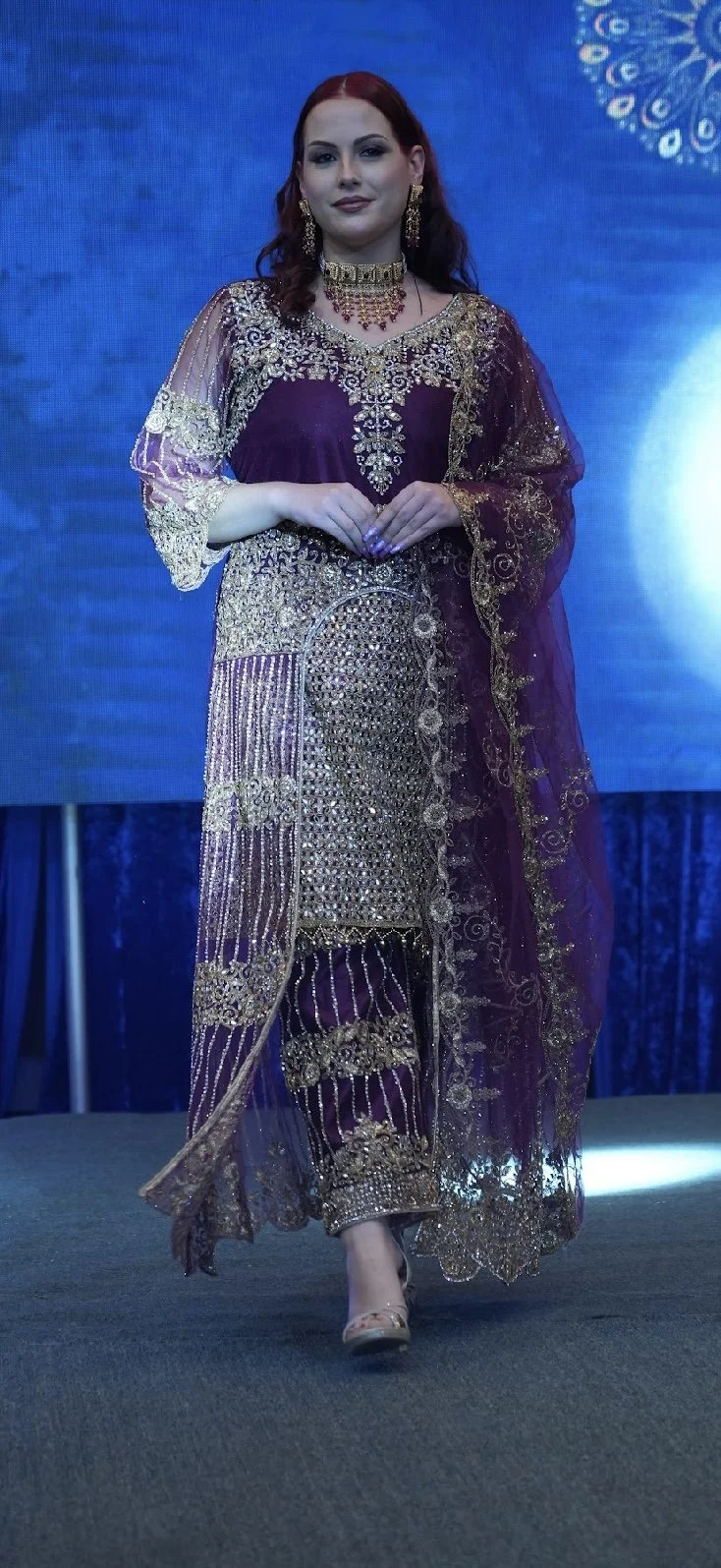A woman walking on stage wearing traditional South Asian attire with gold embroidery and jewelry, against a blue background.