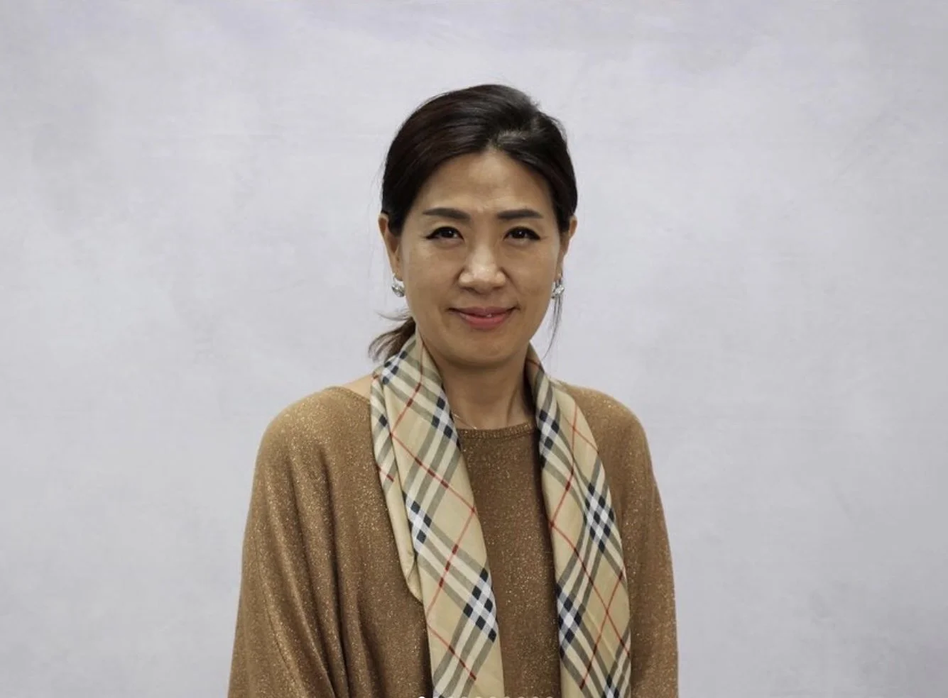 A woman with dark hair in a ponytail, wearing a gold sparkly top and a beige Burberry check scarf, standing in front of a plain white wall.