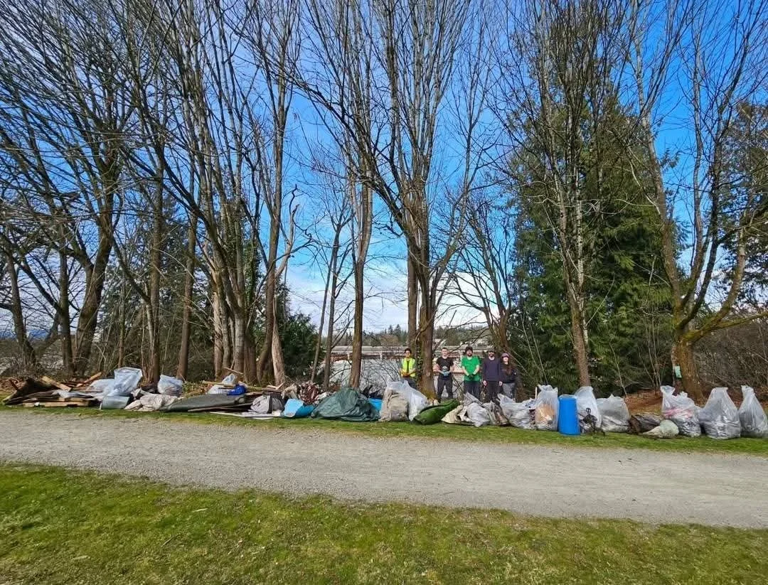 A group of people standing behind a large pile of collected trash and garbage bags beside a wooded area on a clear day.