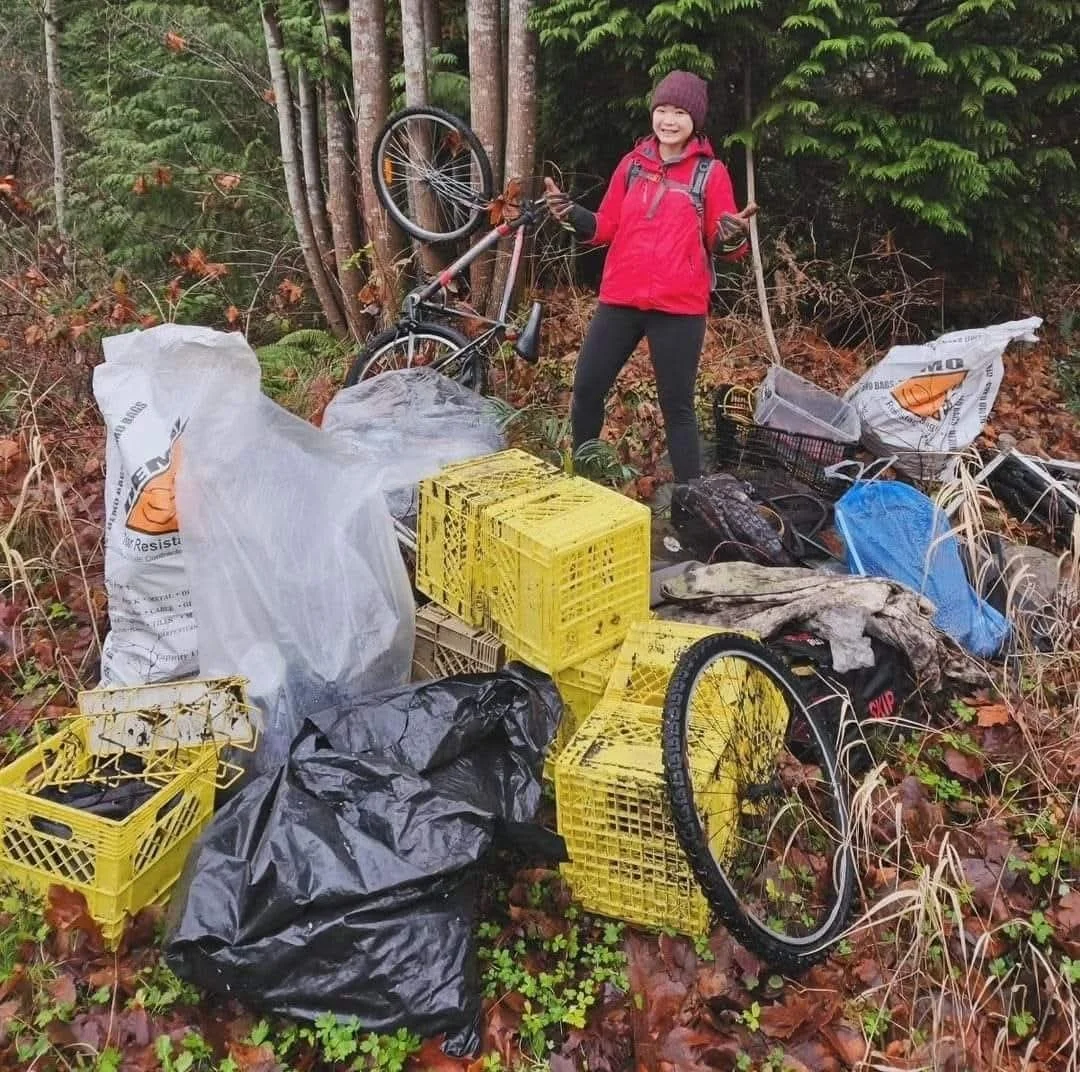 A woman in a red jacket and purple knit hat standing in a forested area next to a bicycle and a pile of trash, including plastic crates, black and white trash bags, various plastic items, and other debris.