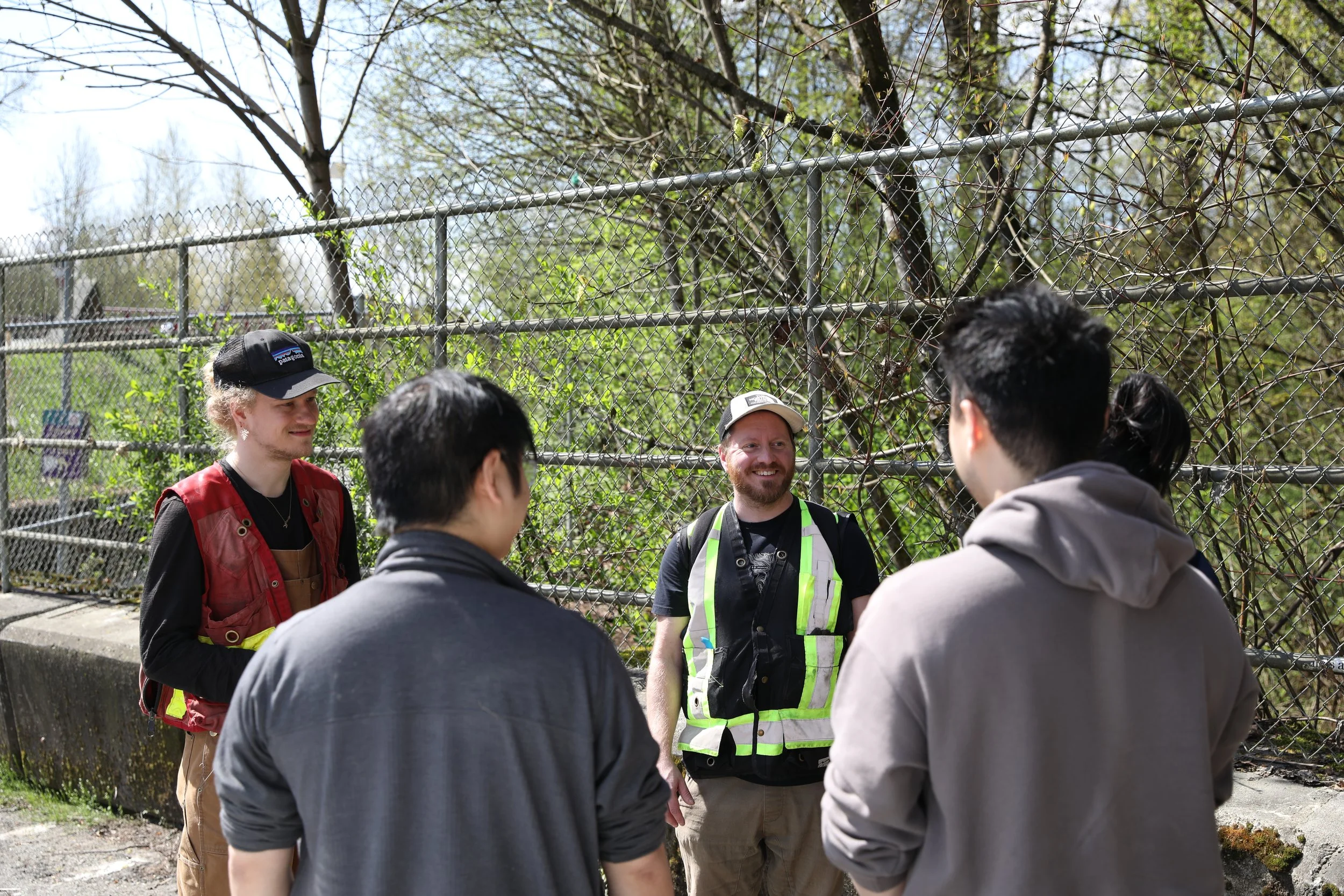 Five people standing outdoors near a chain-link fence, engaged in conversation, with trees and greenery in the background.