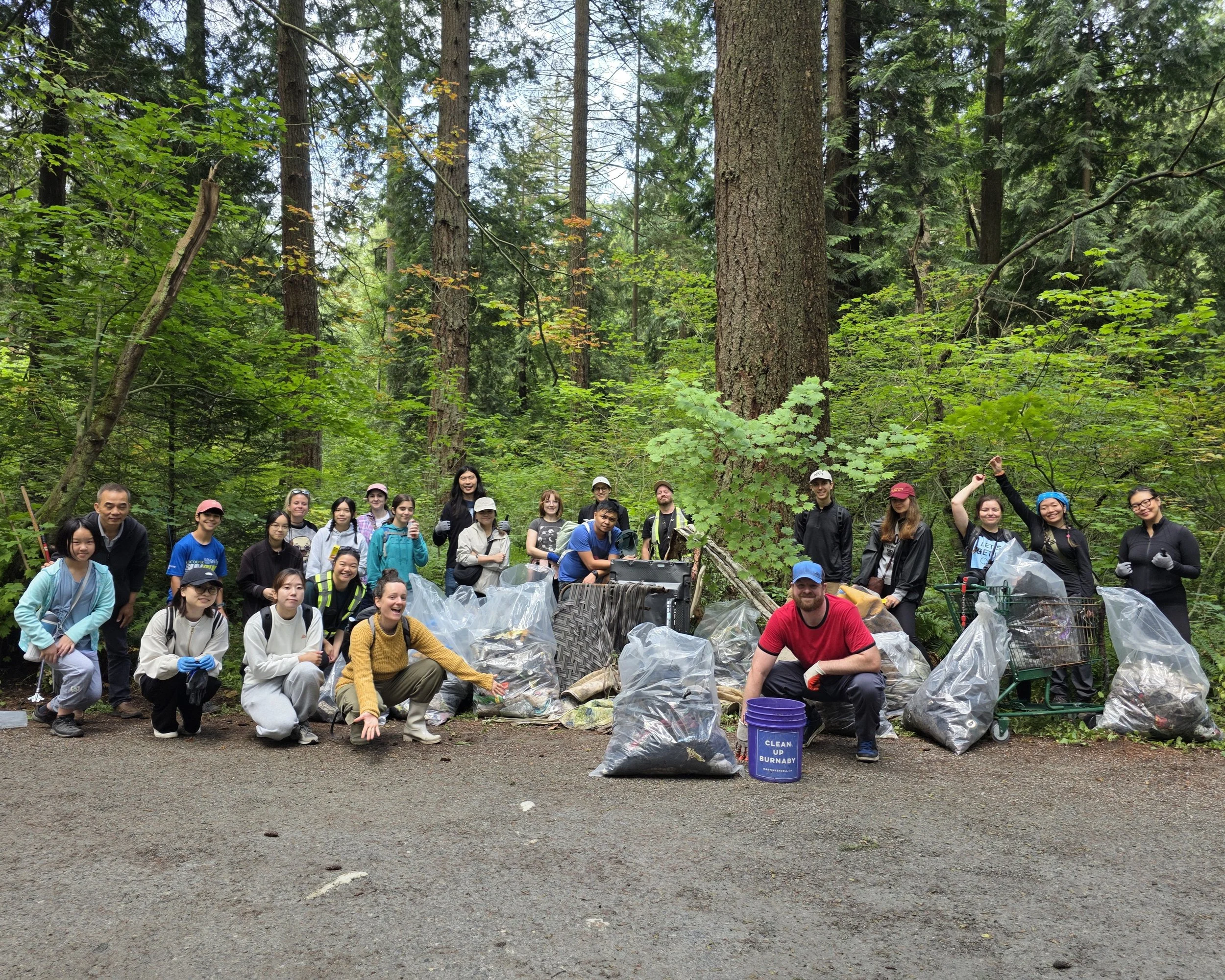 Group of volunteers picking up trash in a forest, surrounded by tall trees and greenery, with bags of collected litter and tools.