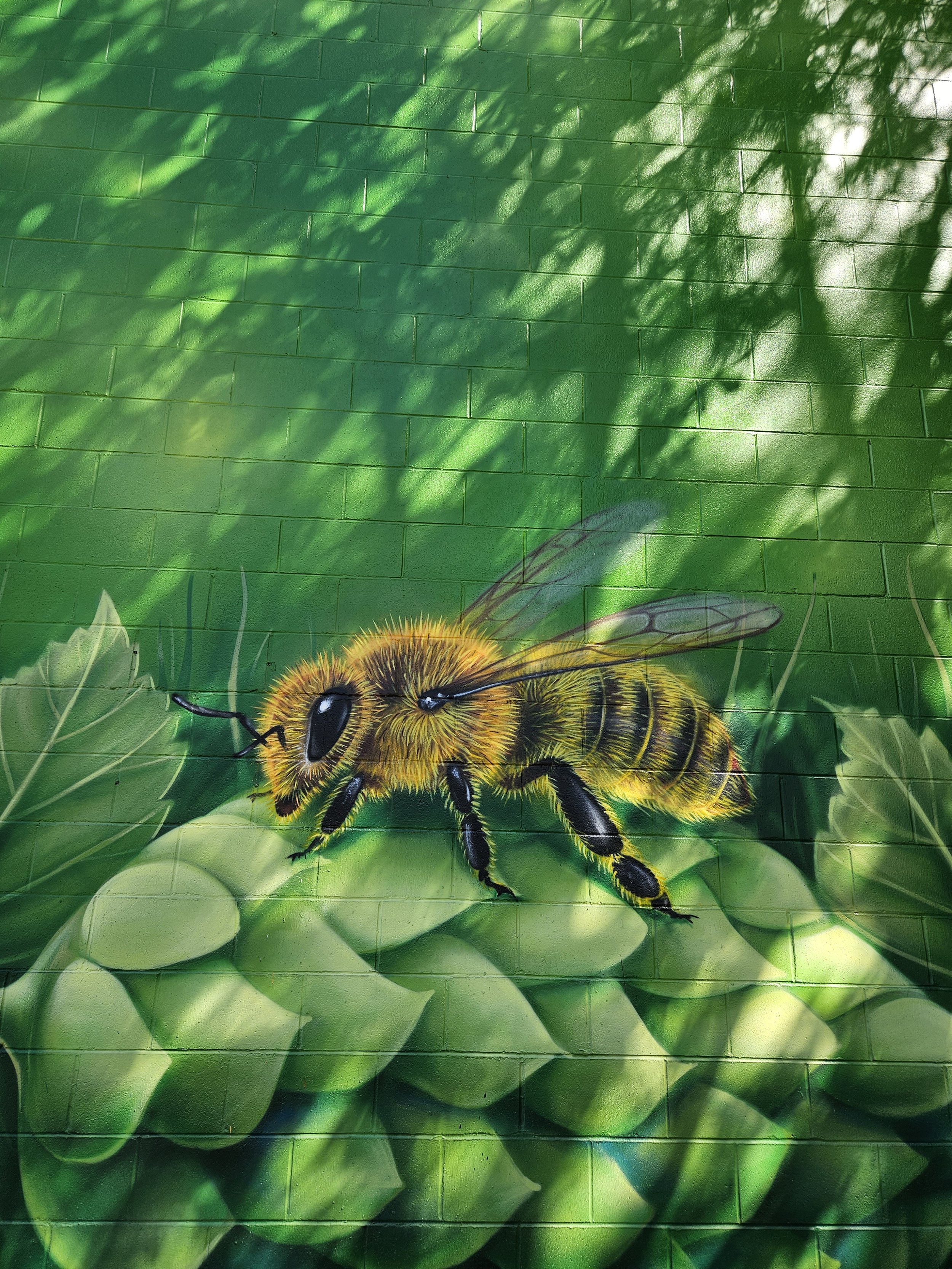 Painted mural of a bee on a green leafy background on a brick wall.