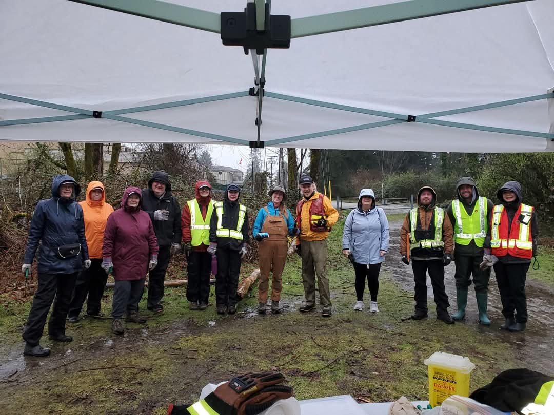Group of people standing outdoors under a white canopy, wearing weather appropriate clothing, some with safety vests and gloves, in muddy terrain with trees and a road in the background.