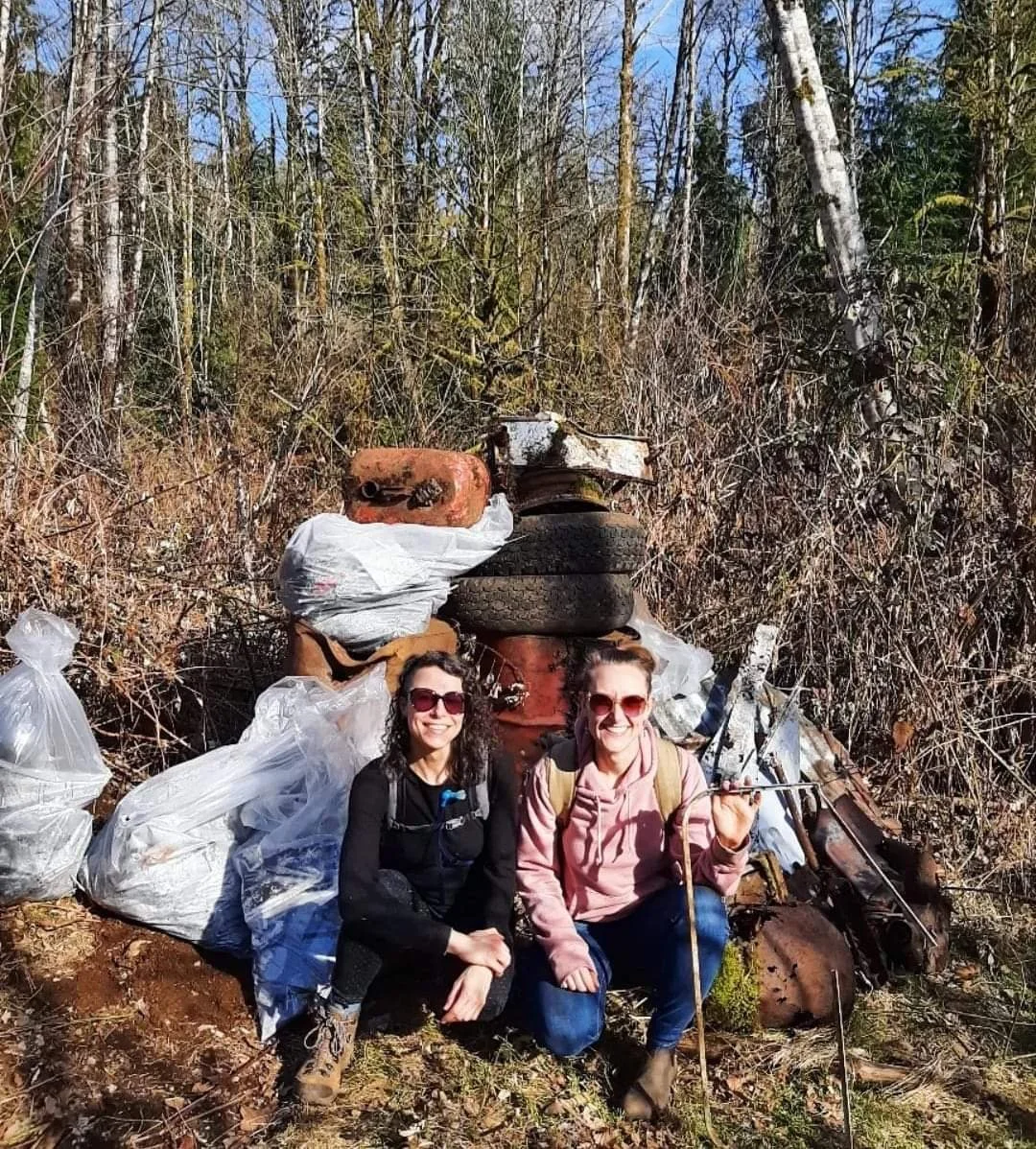 Two women in hiking gear smiling and sitting in front of a pile of trash in a wooded area, including tires, plastic bags, and rusty metal debris.