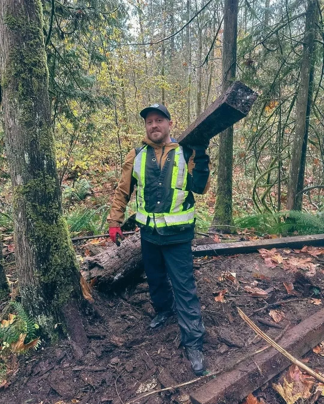A man wearing a high-visibility safety vest, rain jacket, and black pants stands in a forest, holding a large piece of wood above his shoulder. The forest floor is wet and muddy with fallen leaves, and trees surround him.