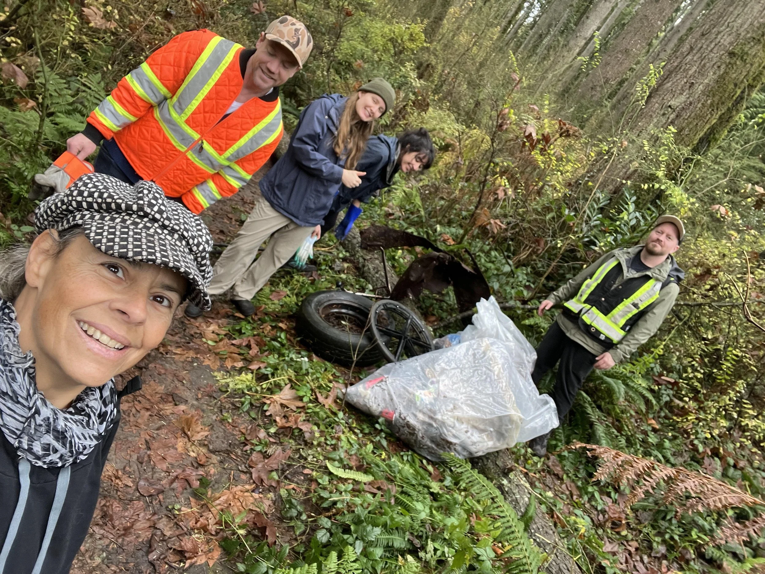 A group of five people cleaning up litter in a forest. Two men and three women, some wearing high-visibility vests and outdoor clothing, are standing near bags of collected trash and discarded tires on the ground.