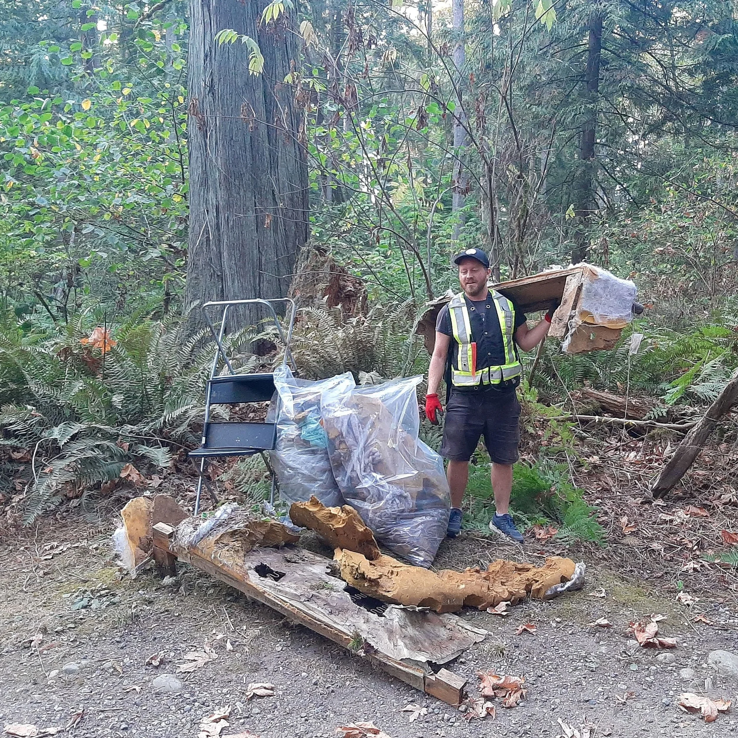 A man in work attire with a safety vest and gloves is carrying a piece of wood in a forested area. There is debris and trash scattered on the ground around him, including broken wood and a plastic bag, indicating cleanup or removal activity.