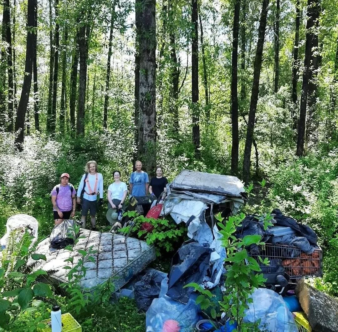 Group of five people in a forest next to a large pile of trash and discarded items.