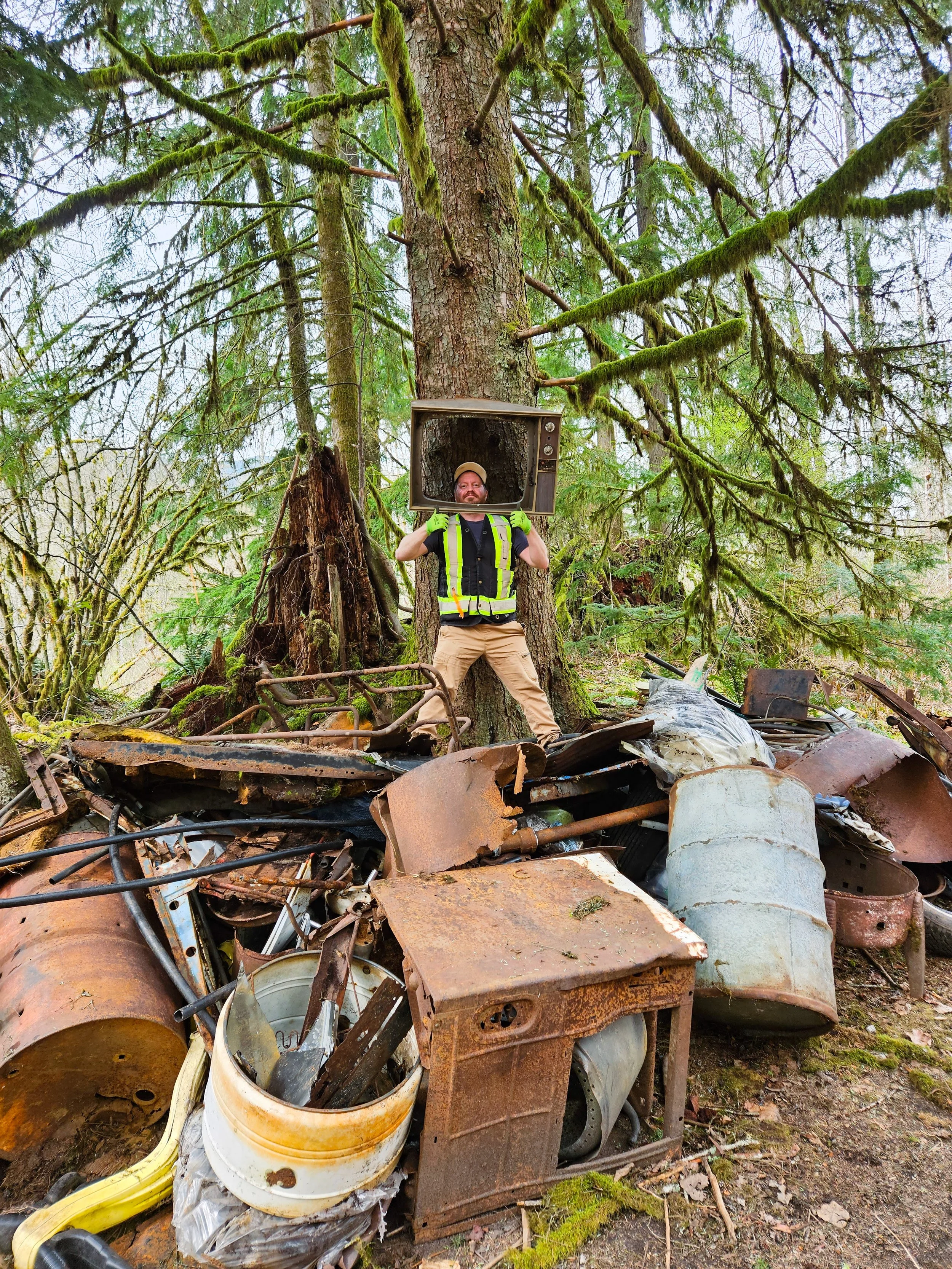 A man wearing a safety vest and khaki pants standing among rusted metal debris in a forest, holding an old television set on his shoulders, with tall green trees and moss in the background.