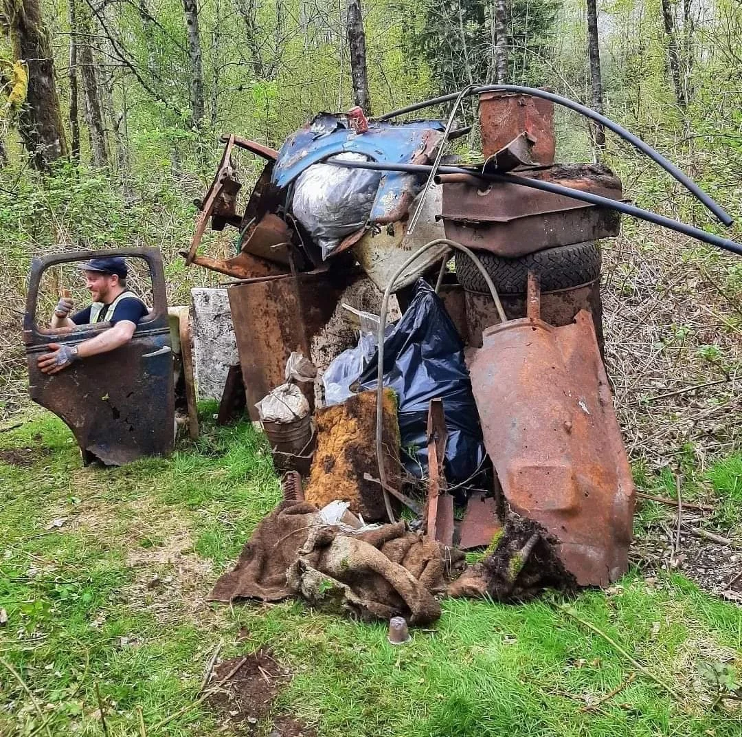 A pile of rusted, old scrap metal includes various car parts like a door, tires, and other unidentifiable metal pieces, situated in a grassy area in a wooded forest.