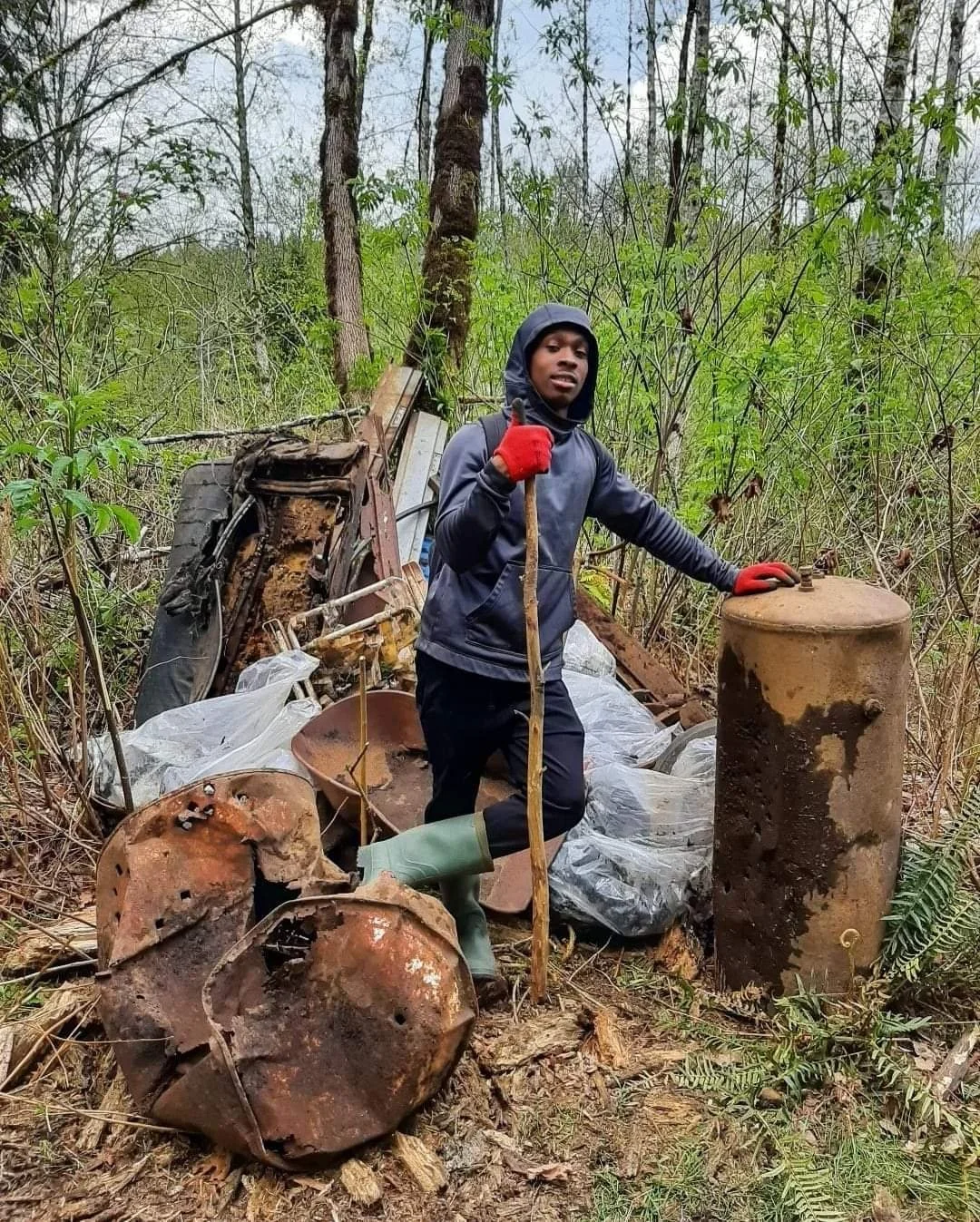A young man wearing a dark hoodie, red gloves, and rubber boots standing in a wooded area with discarded rusty metal objects and debris around him, holding a stick.