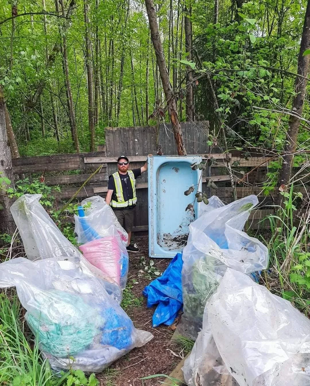 A man in a black shirt, sunglasses, and a reflective safety vest stands next to a discarded blue mini refrigerator in a wooded area. Several large plastic trash bags filled with garbage are scattered around the ground.