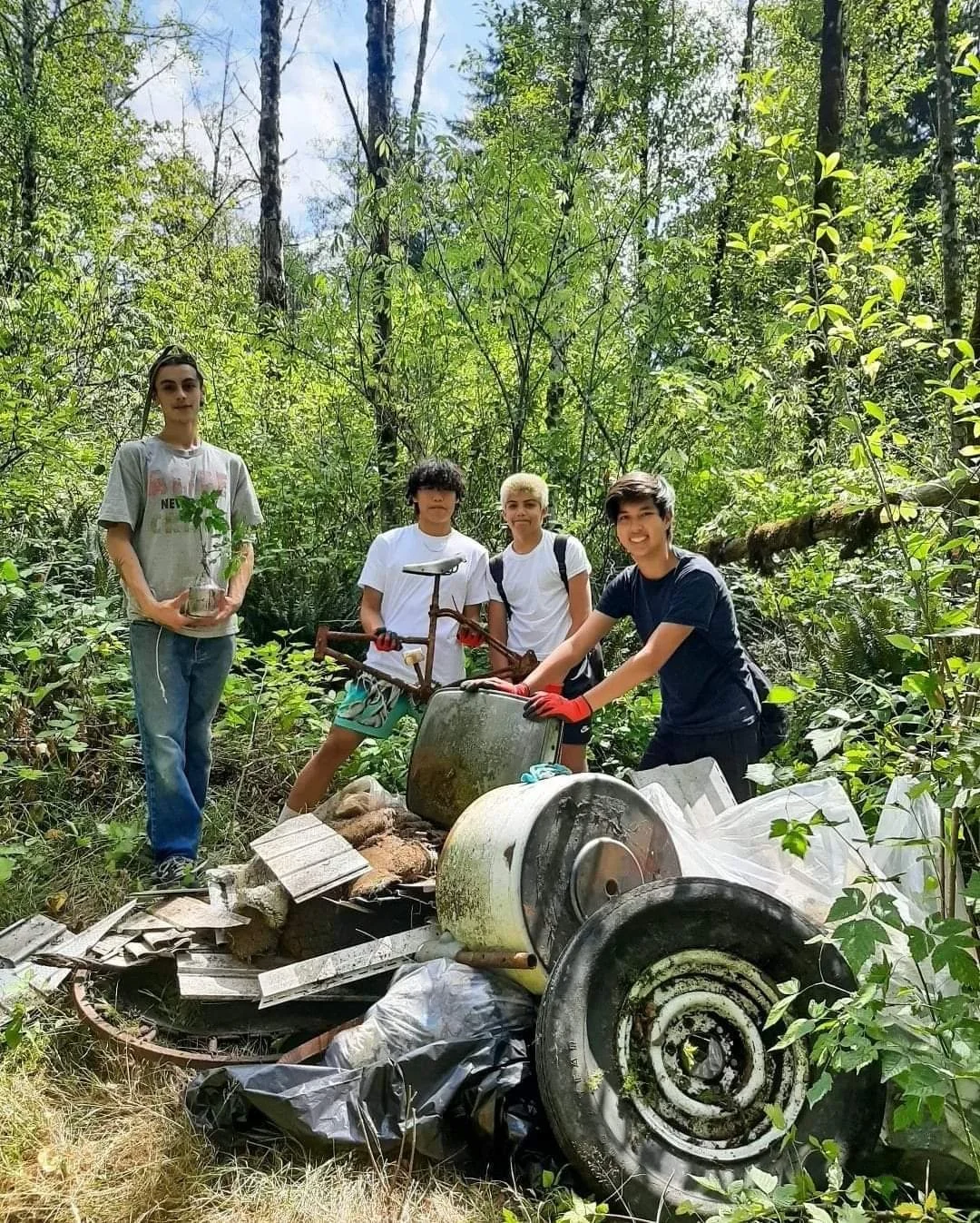 Five young people cleaning up trash in a forest, with debris including a large metal can, plastic bags, broken metal parts, and an old bicycle.