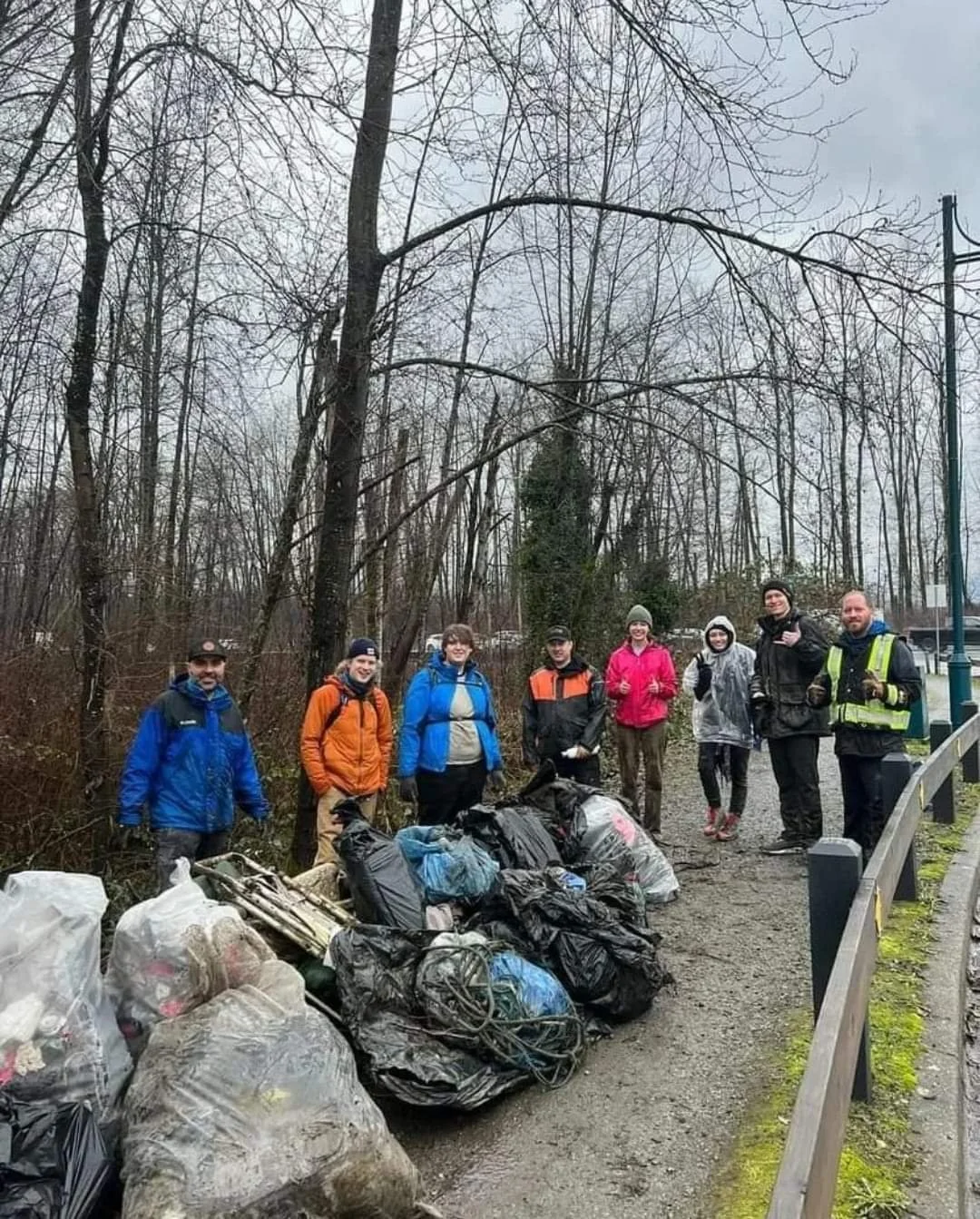 Group of people participating in a cleanup event outdoors, standing next to bags of collected trash on a dirt path in a wooded area on a cloudy day.