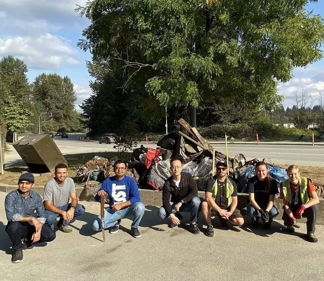 Group of seven people kneeling and sitting in front of a large pile of trash and debris on a sidewalk, with a tree and road with cars in the background.