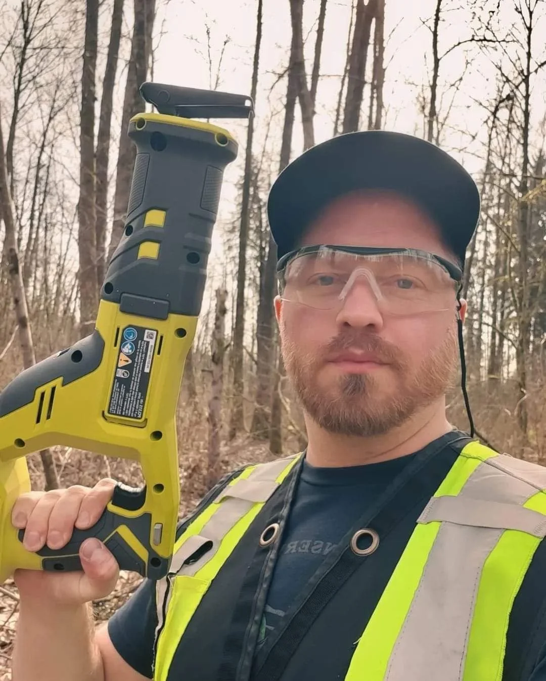A man wearing safety glasses, a baseball cap, and a reflective safety vest holding a cordless power tool in a wooded area during daytime.