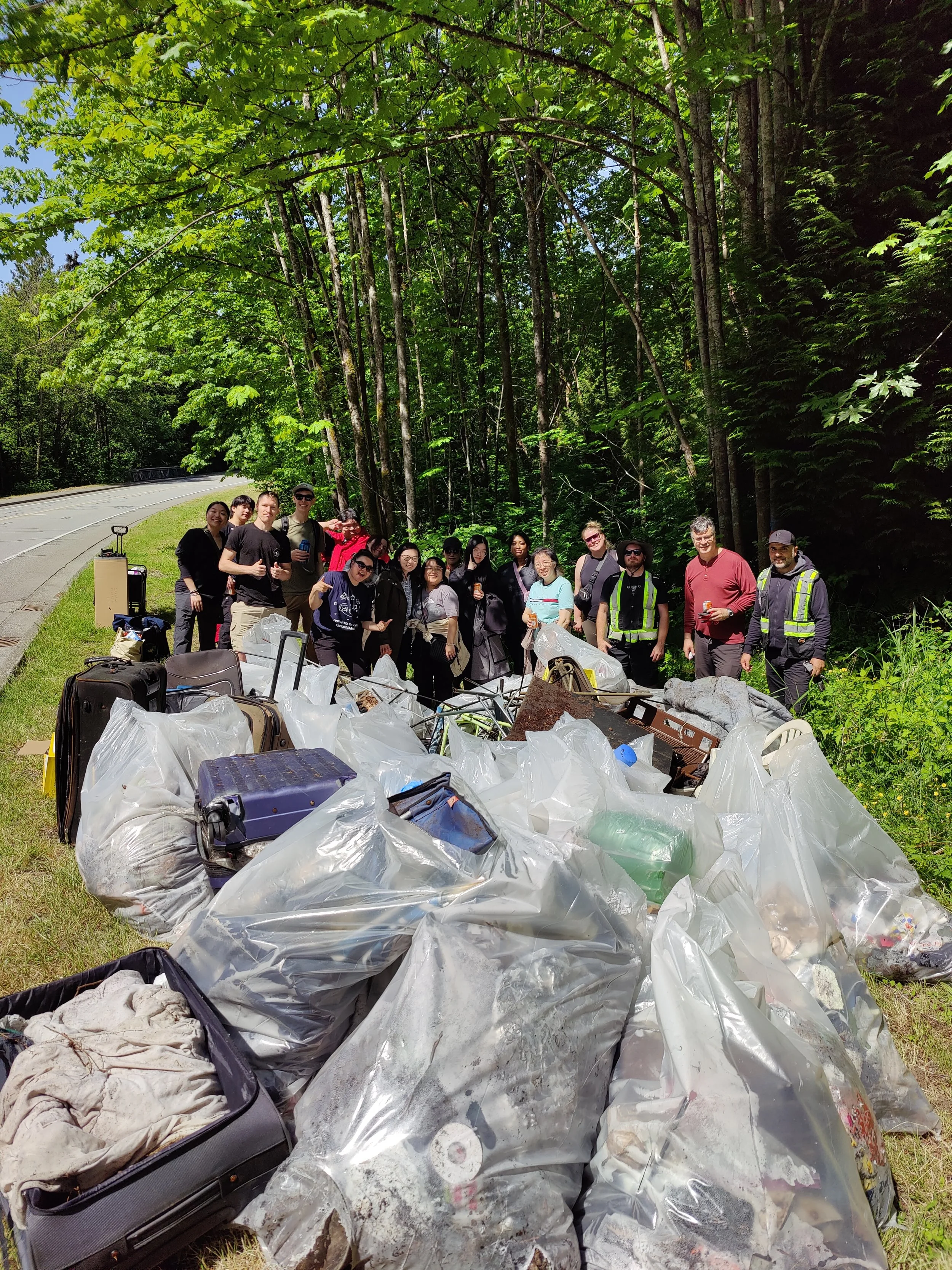 Group of people standing behind a pile of trash and garbage bags on the side of a forested road, after a cleanup event.