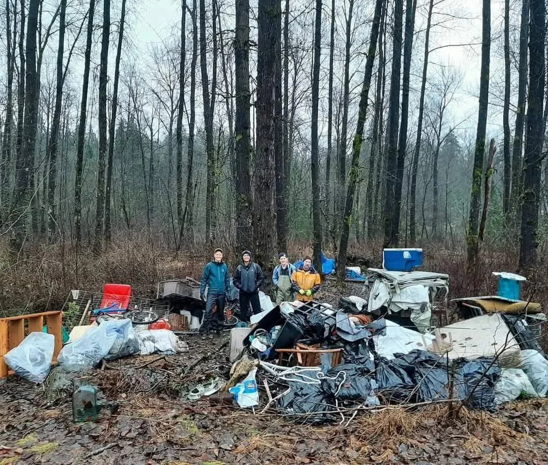 Four people standing in a forest surrounded by trash and debris, including furniture, bags, and miscellaneous objects.