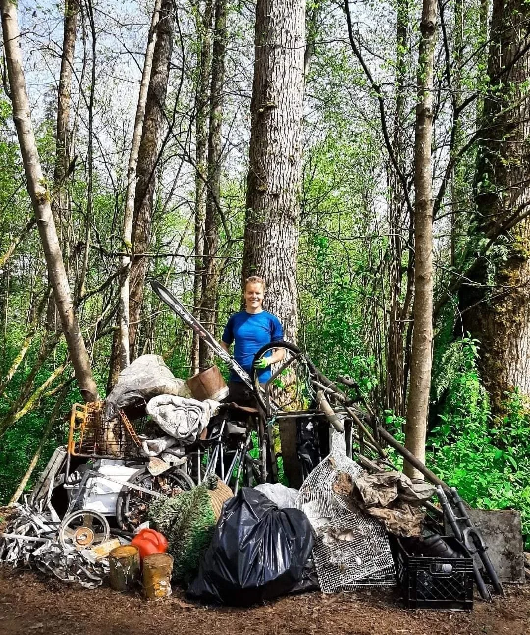 A person in a blue shirt standing behind a large pile of trash and discarded household items in a forested area.