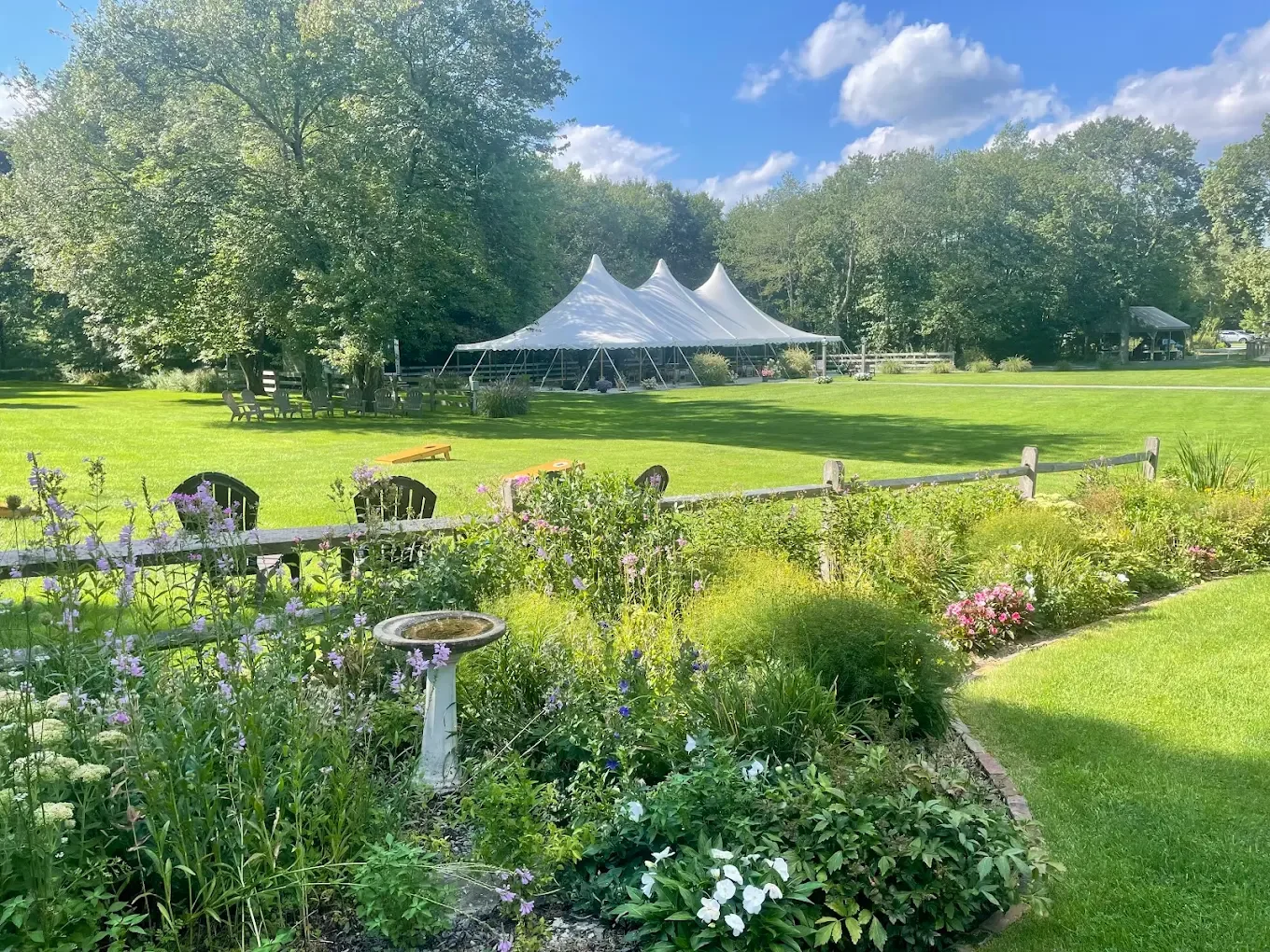 A lush green outdoor space with a white event tent, surrounded by trees, colorful flowers, and garden furniture under a blue sky with scattered clouds.