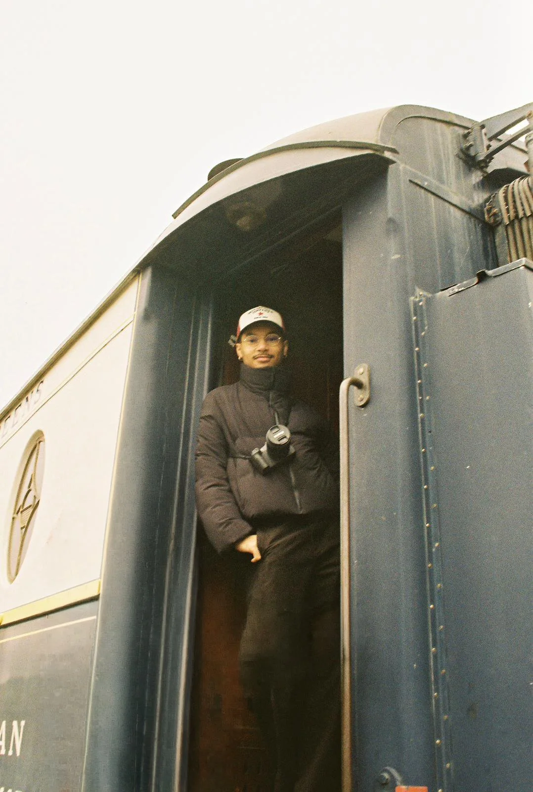 Personne debout dans le cadre d'une porte de train à vapeur, portant une veste noire, un chapeau blanc, et une caméra autour du cou.