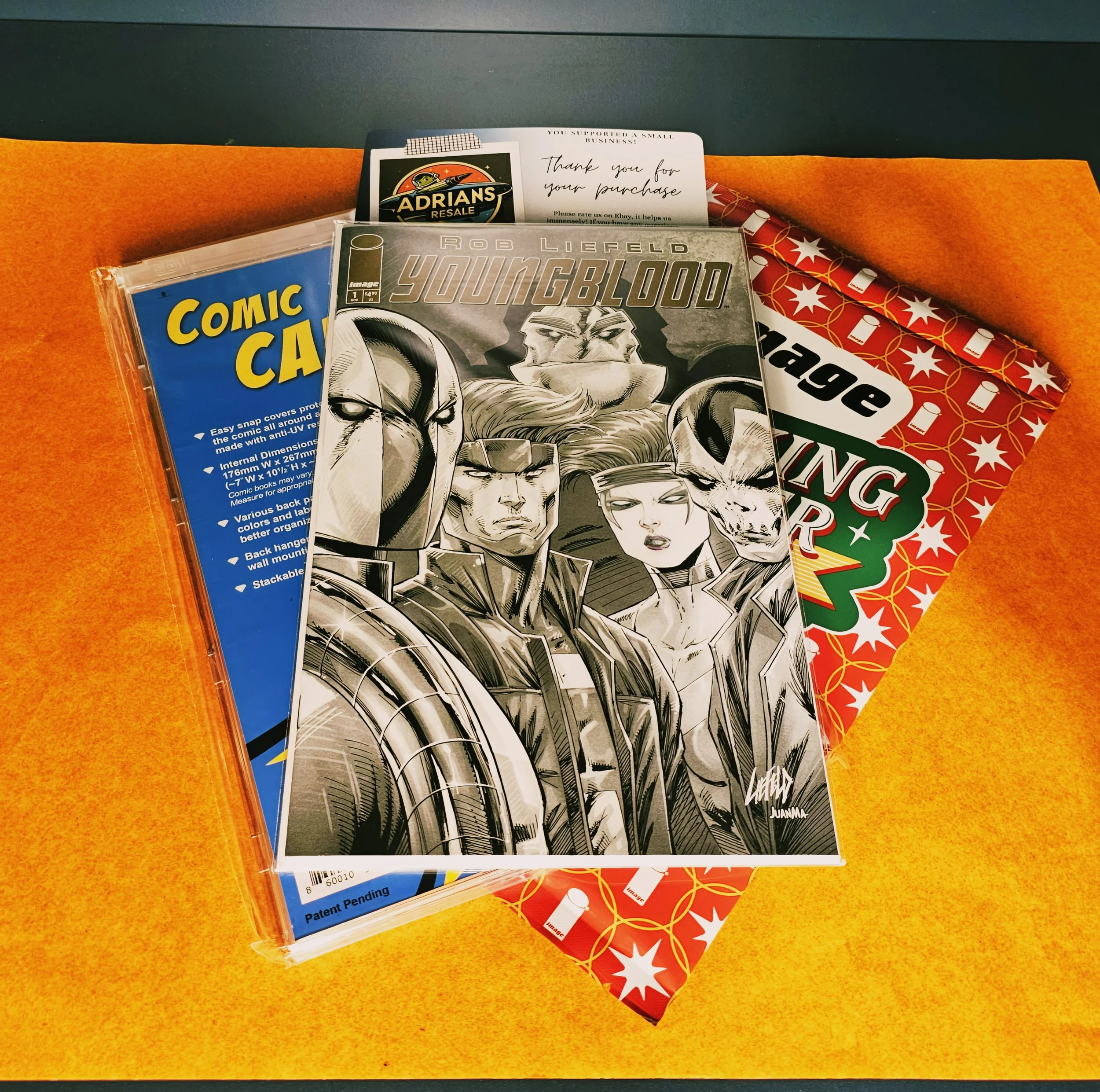 A comic book titled 'Youngblood' by Rob Liefeld resting on a Christmas gift bag with red and white star pattern, and Christmas lights. Part of a gift package with a note expressing thanks for a purchase.