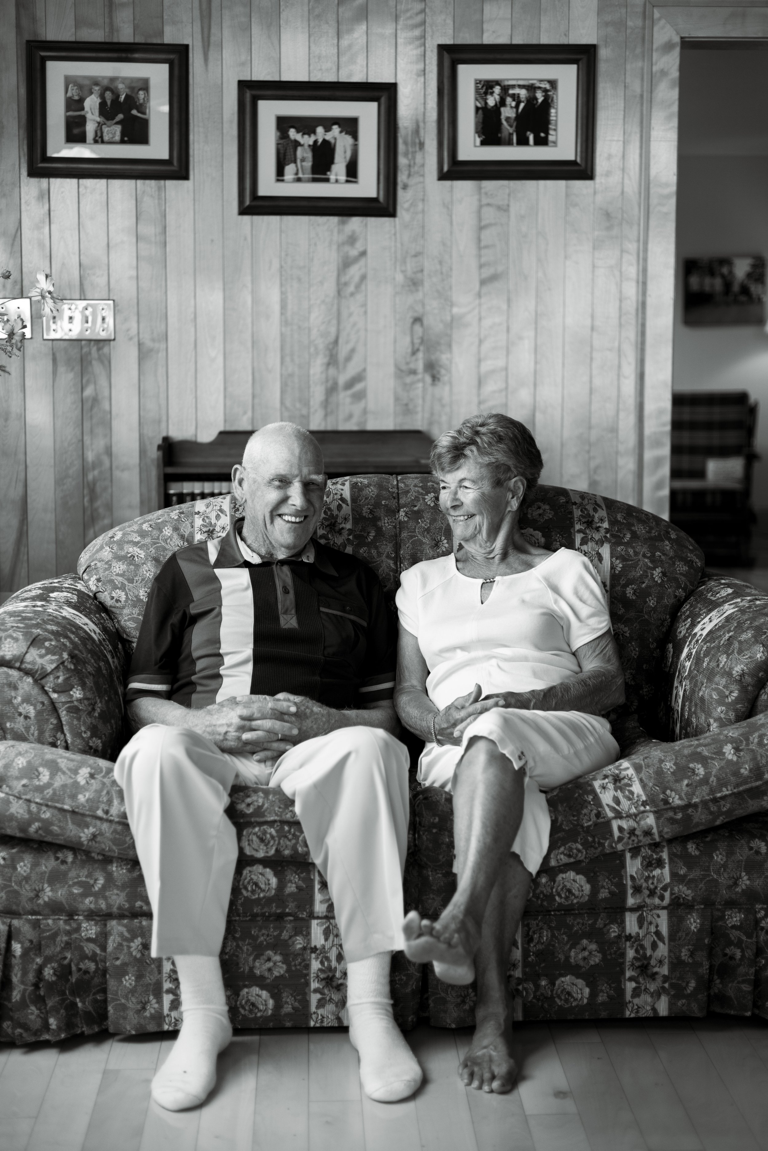 An older couple sitting on a floral patterned couch in a room with wooden wall paneling, laughing and looking at each other, with framed photographs hanging on the wall behind them.
