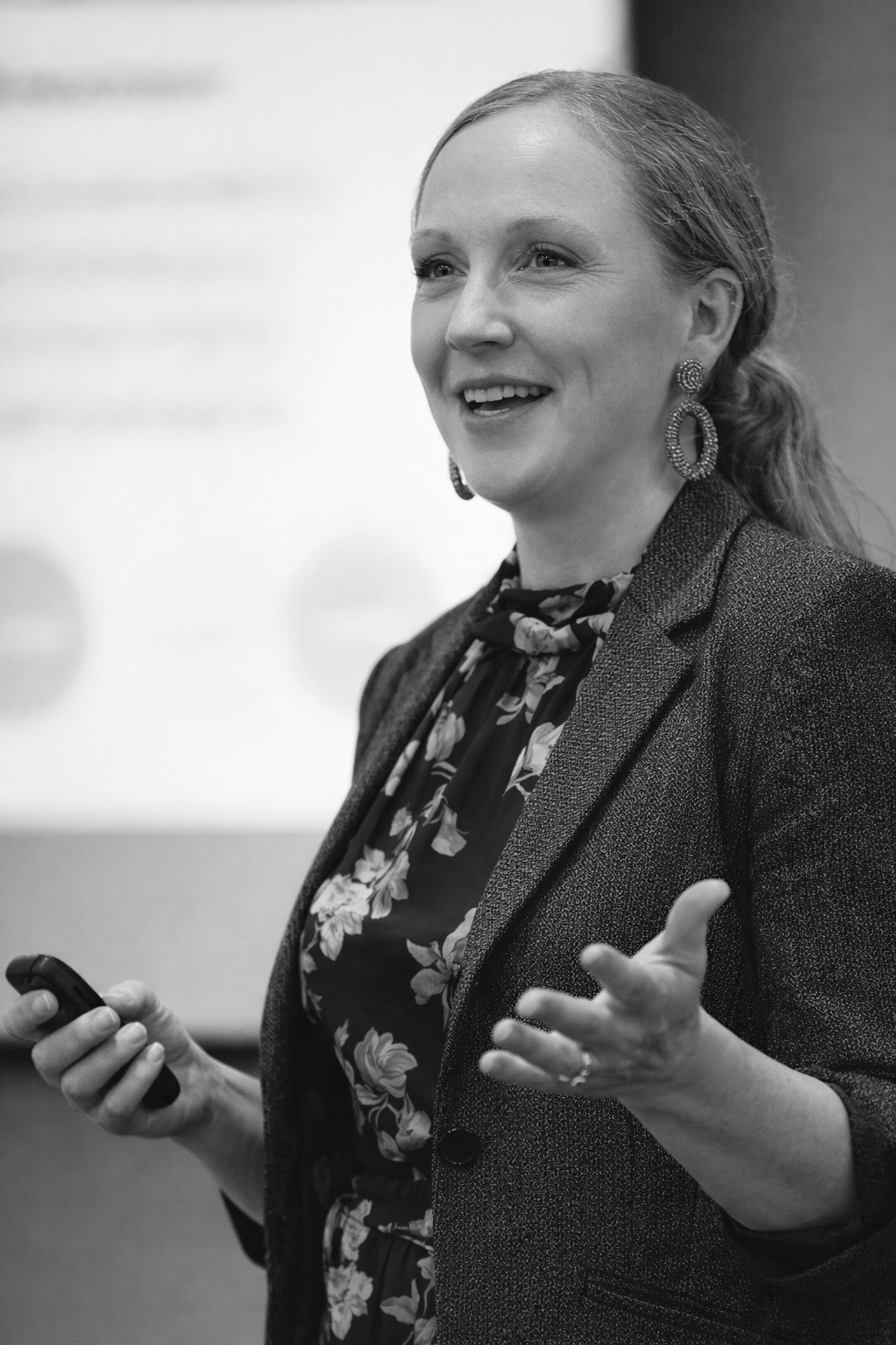 Black and white photo of a woman giving a presentation, holding a remote in her right hand and gesturing with her left, dressed in a blazer with a floral blouse and large earrings.