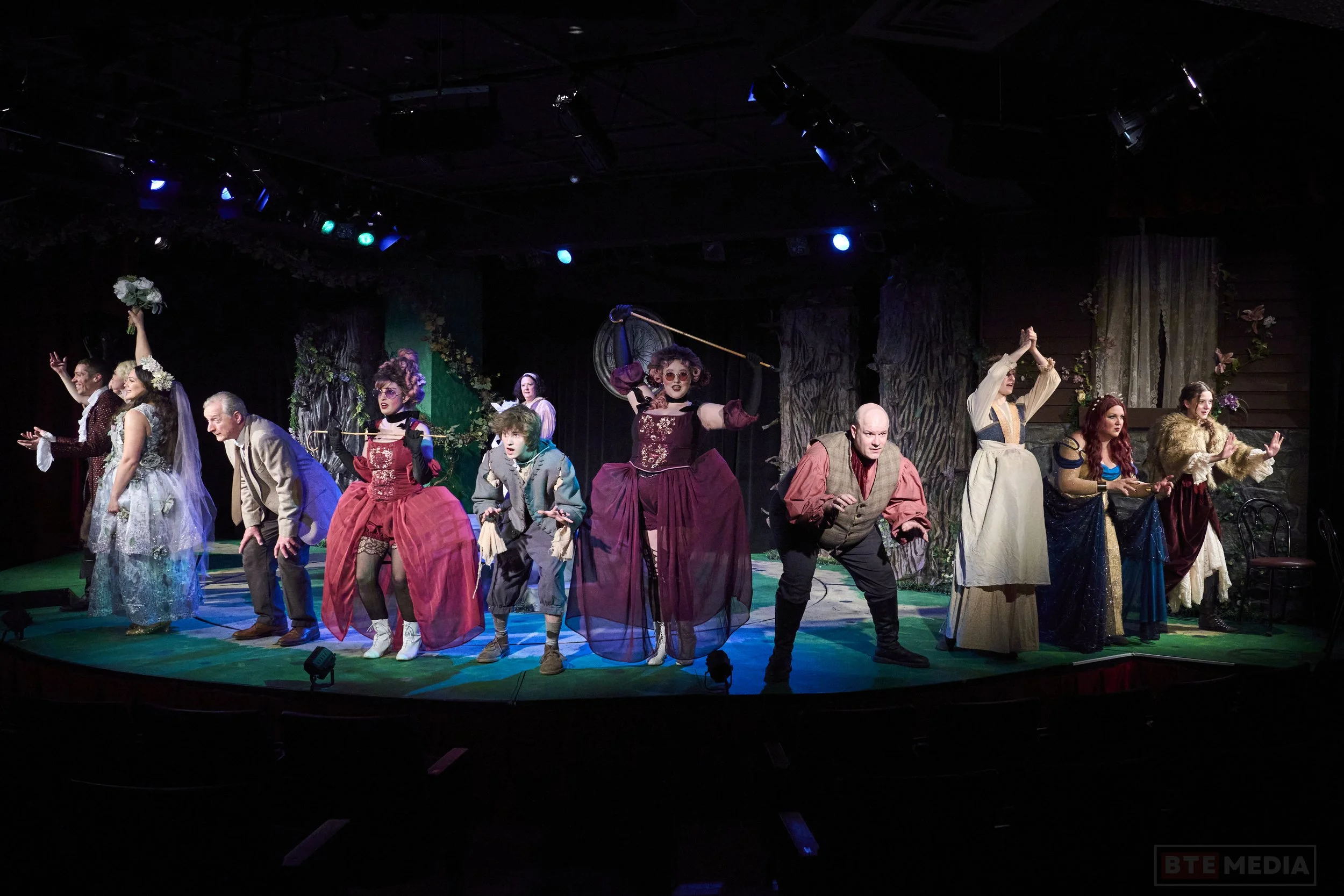 Theater cast performing on stage in period costumes with forest scenery backdrop, characters in expressive poses.