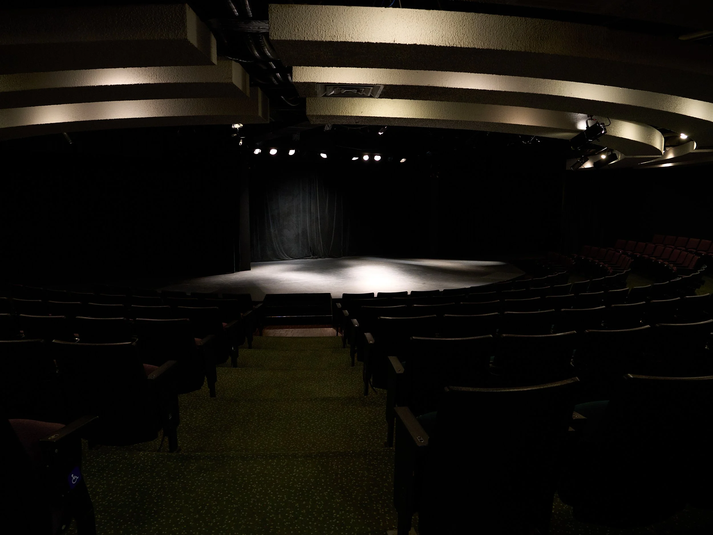 Empty theater with a stage and black curtains, illuminated by overhead lights, with rows of dark seats.