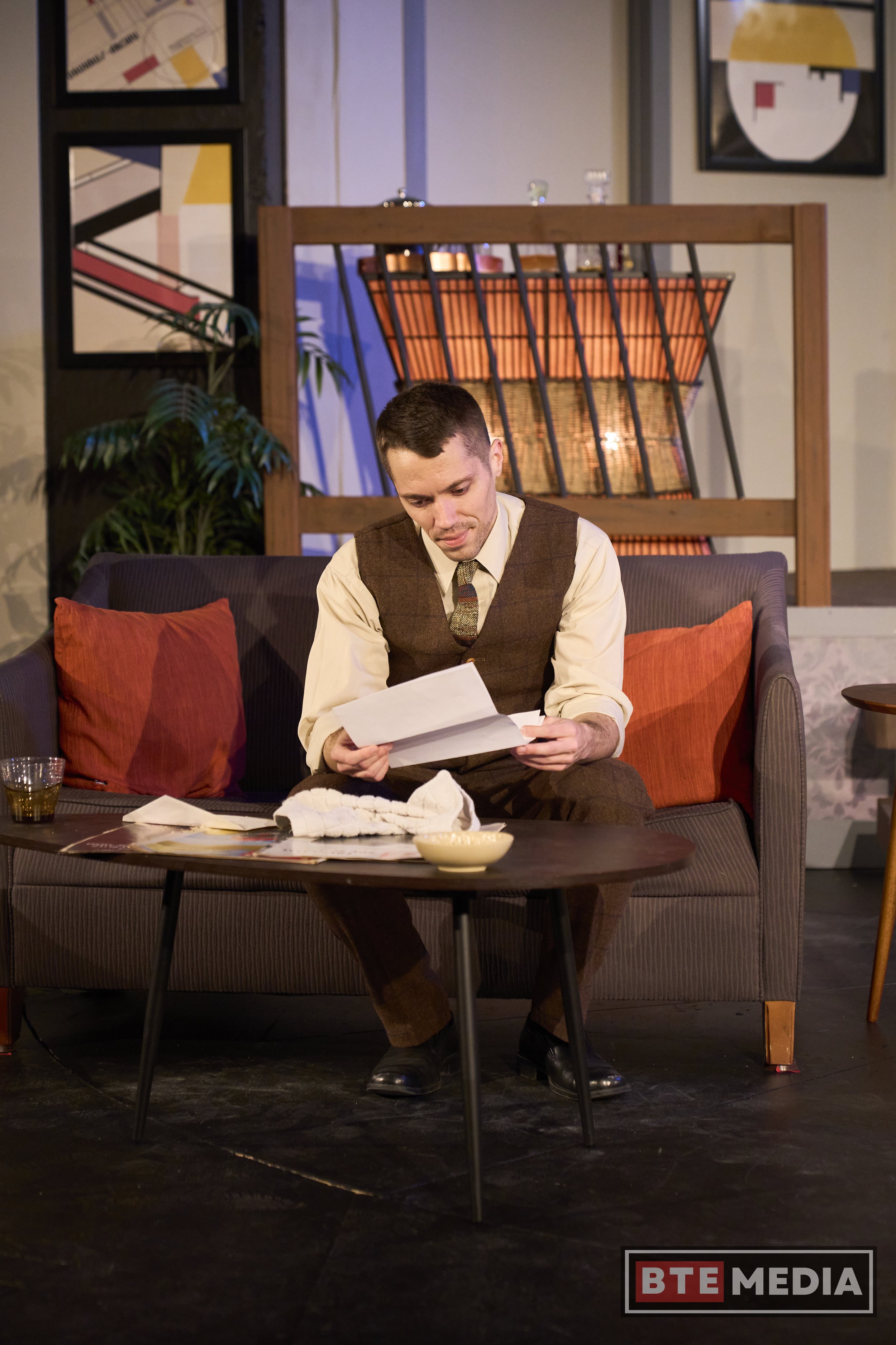 A man sitting on a sofa, looking at papers, in a living room or stage set with colorful abstract artwork on the walls and a wooden bookcase behind him.