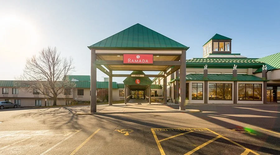 Exterior view of a Ramada hotel with a green roof, a covered entrance, and parking lot in front, during daytime.