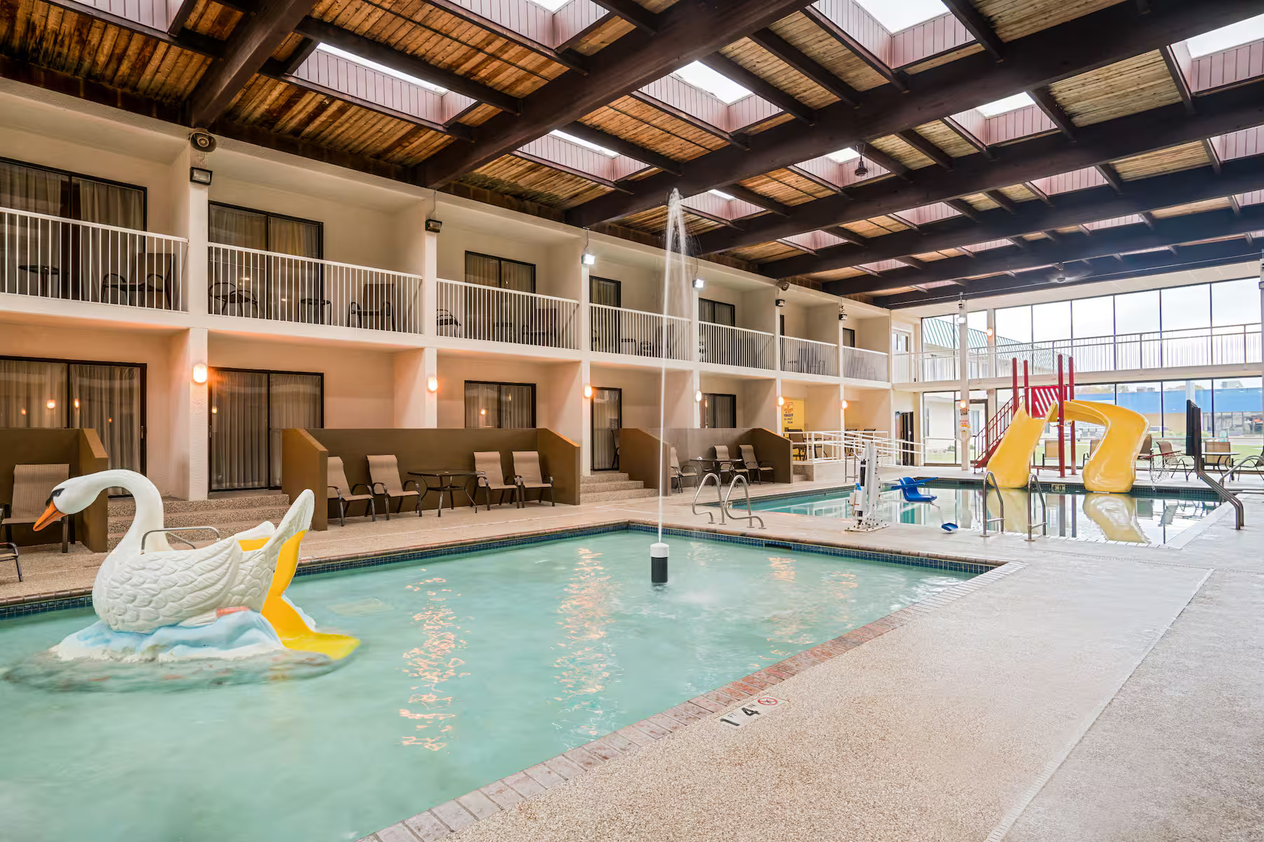 Indoor pool area with a swan-shaped float, water slide, and a small fountain feature. Pool chairs are along the side, and a second level balcony surrounds the area.
