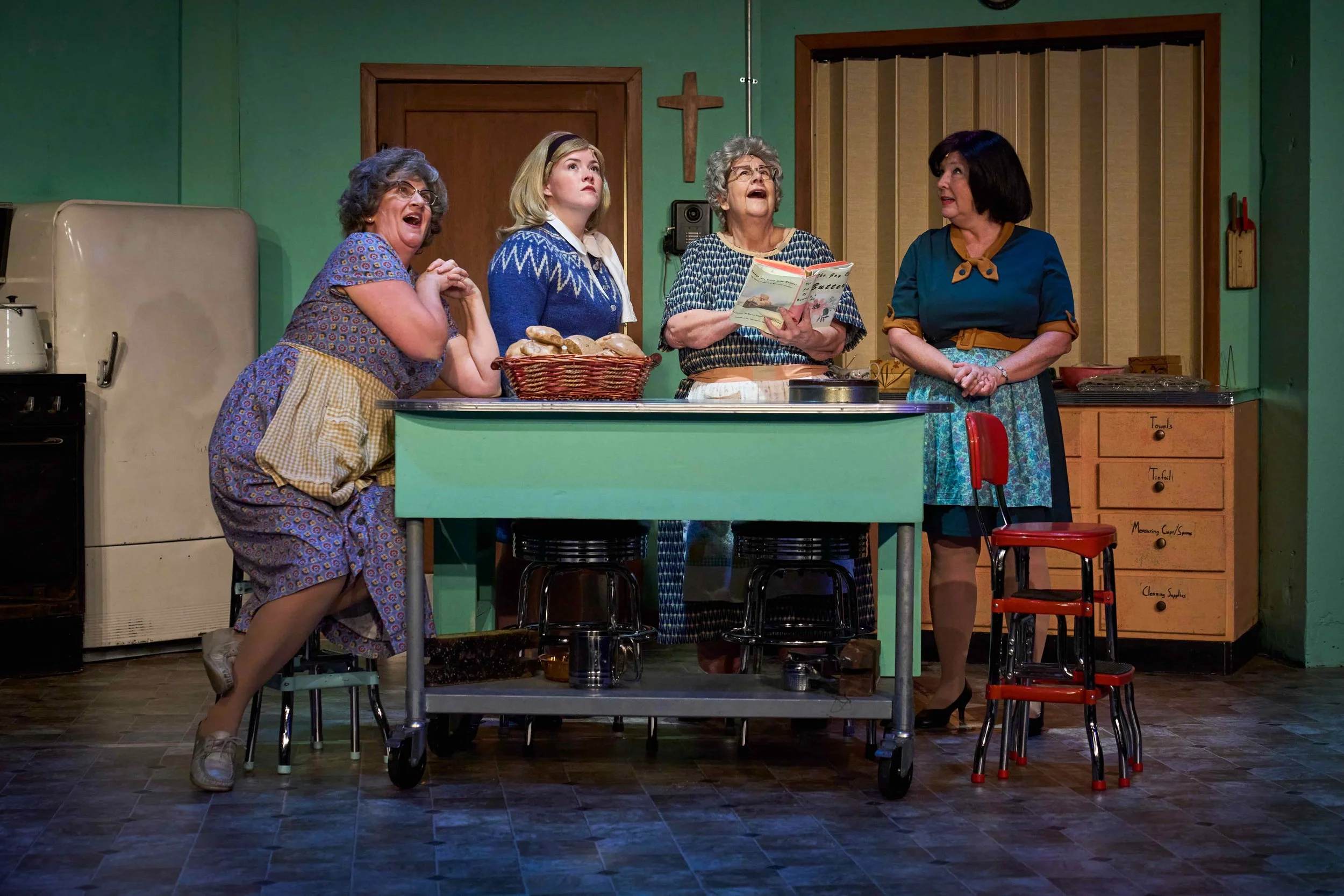 Four women dressed in vintage clothing standing around a kitchen counter, with three of them singing or performing, and one holding a cookbook, in a retro kitchen setting.