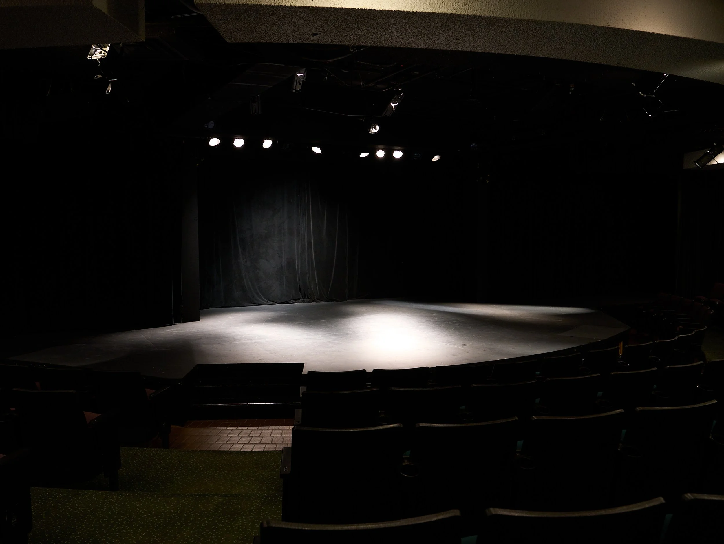 Empty theater stage with focused lights, black curtains, and seating area in front.