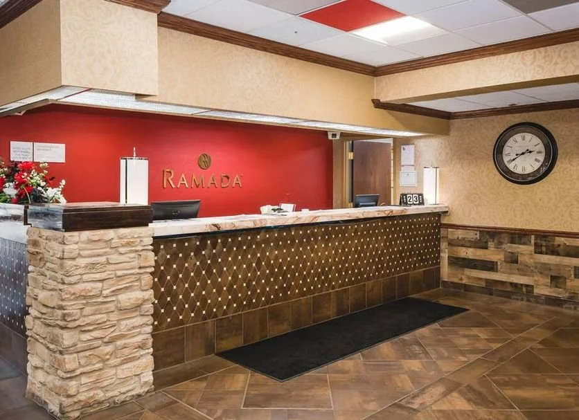 Hotel reception desk with a red wall backdrop, a sign that says 'Ramada,' a black watch on the wall, and a flower arrangement on the counter.
