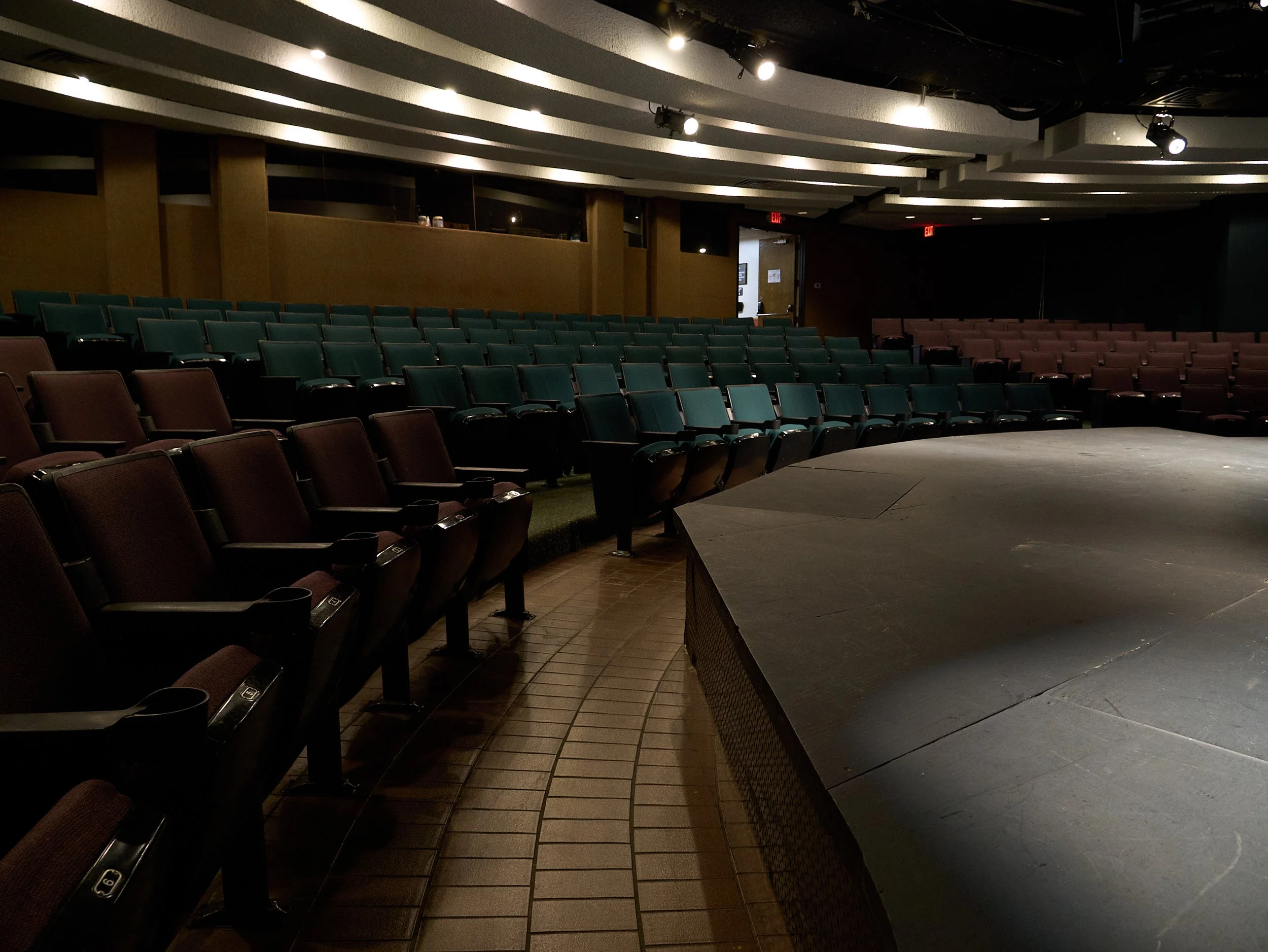 Empty theater with stacked chairs and a stage, dim lighting, wooden floor and exit signs.