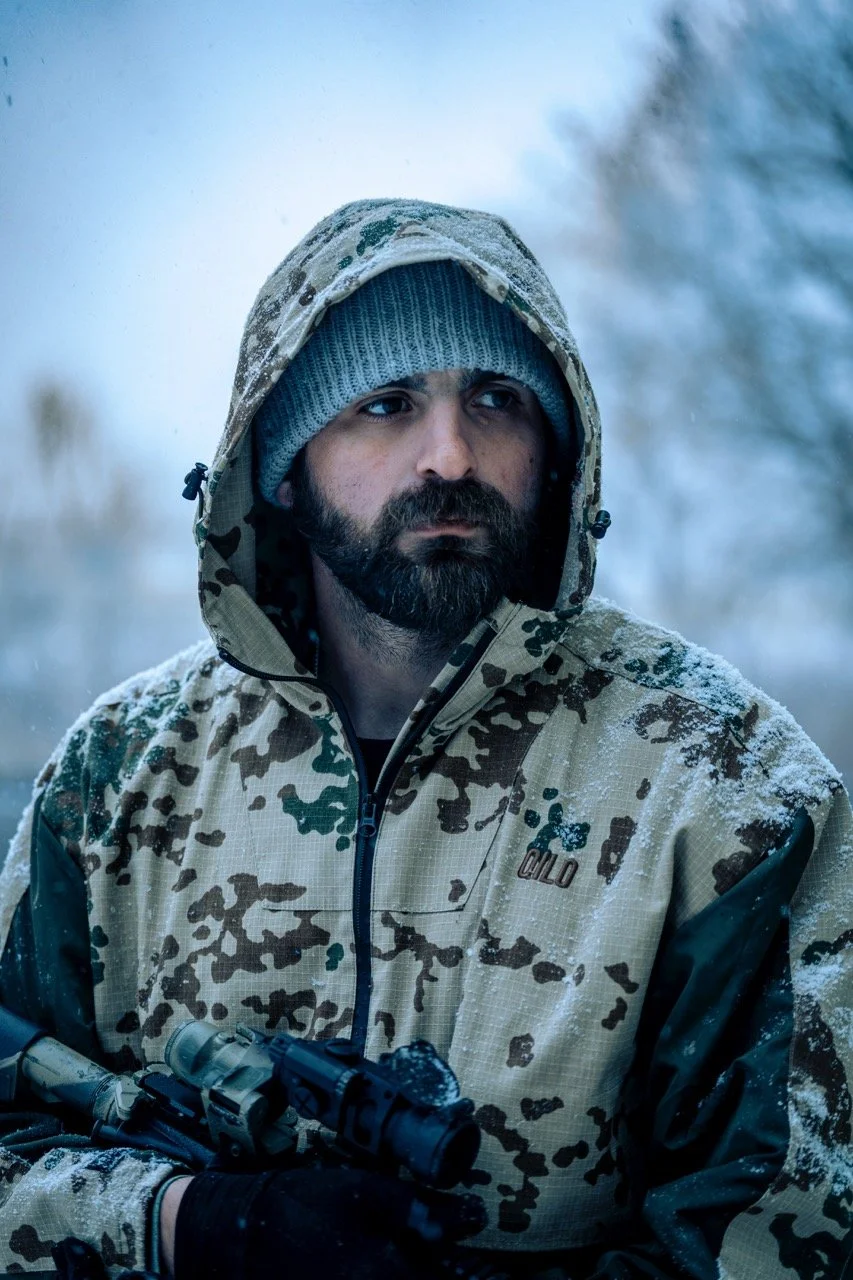 A man with a beard wearing a camouflage jacket, gray beanie, and hood, holding a rifle, outdoors in a snowy environment.