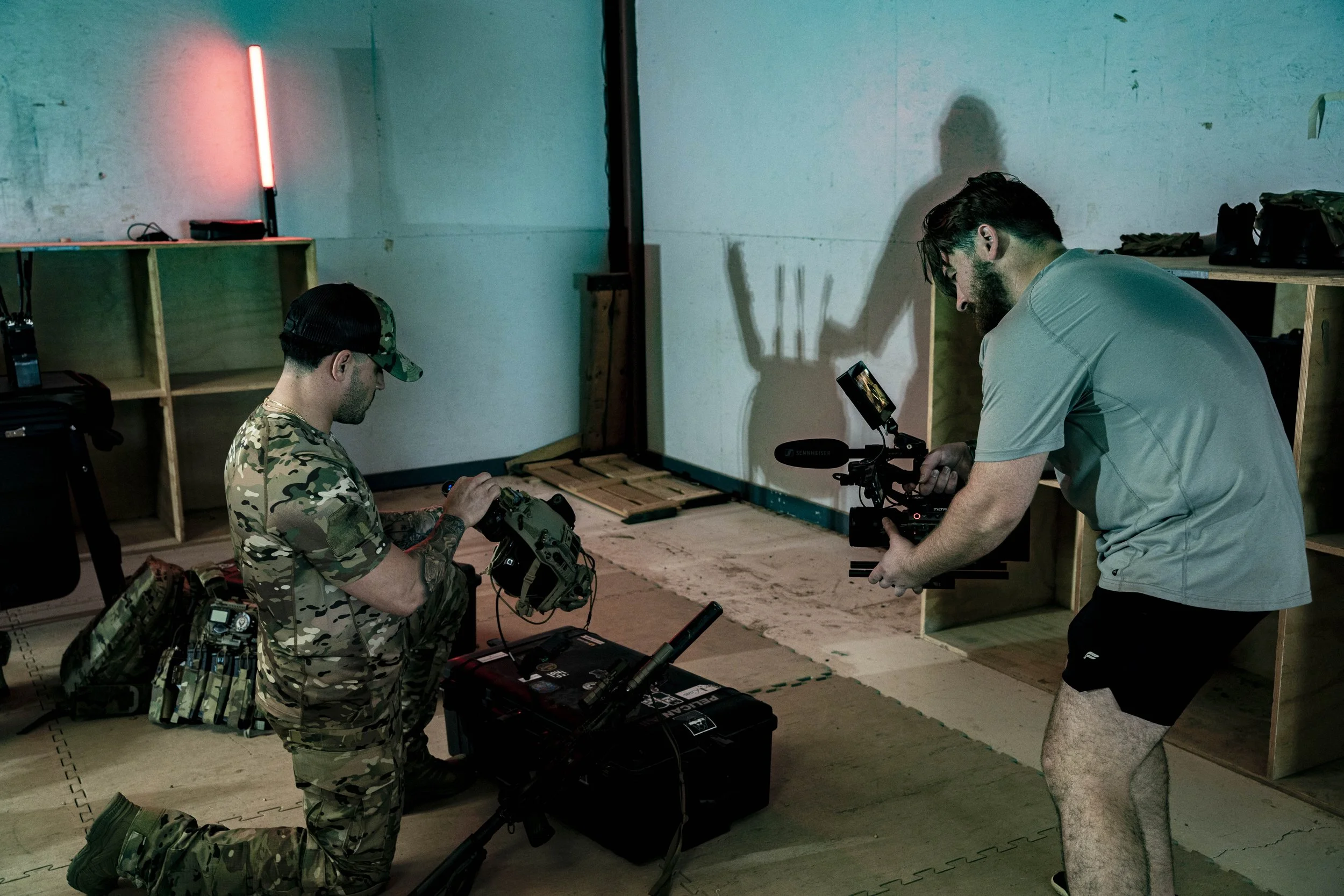 Two men are filming in an indoor setting with cubed shelves; one is in military camouflage kneeling and working with equipment, the other in a gray T-shirt and shorts operating a camera. There's a neon light on the wall, and the room has a plain appe