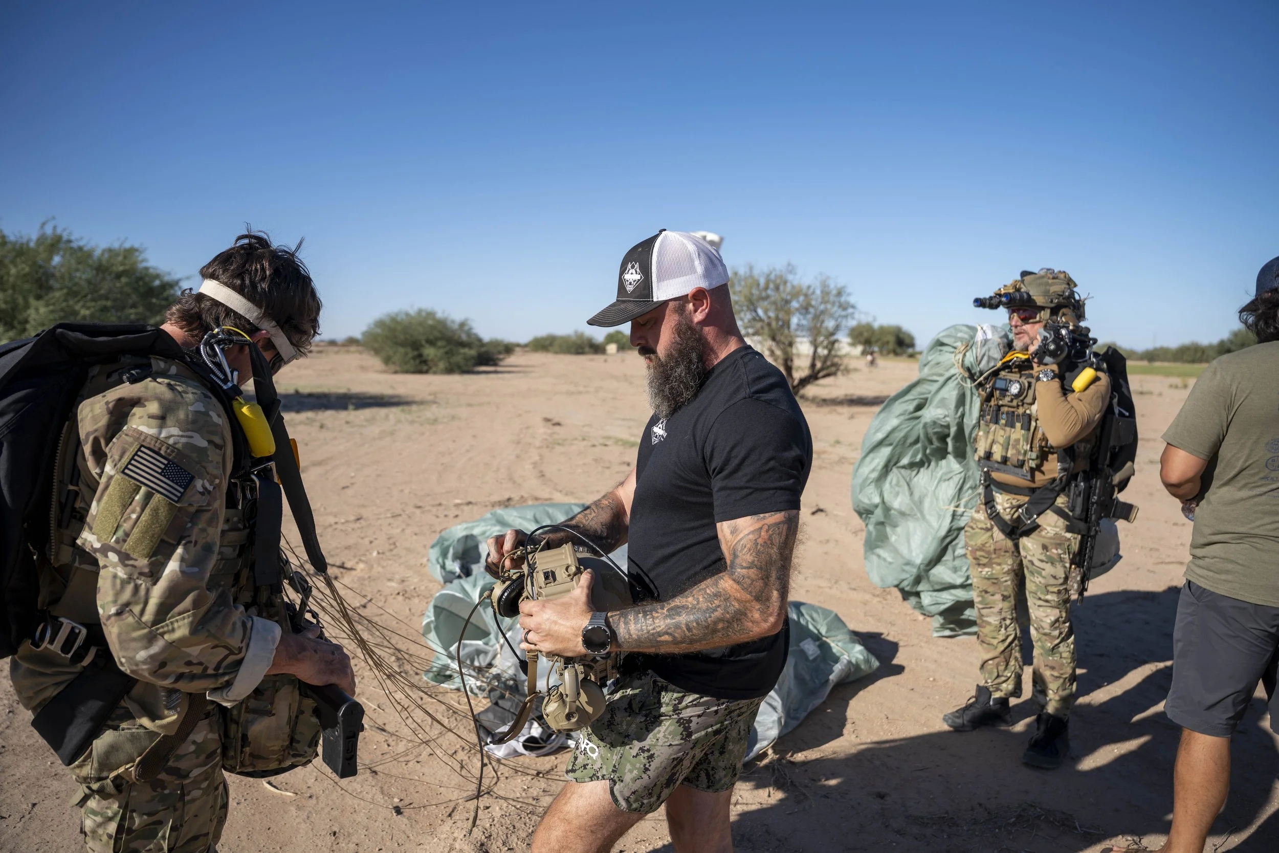 Four people in desert outdoor setting, two are dressed in military gear with backpacks and tactical equipment, one holding a camera, one in camouflage shorts and black t-shirt, others in green shirts, with clear blue sky and sparse trees in backgroun