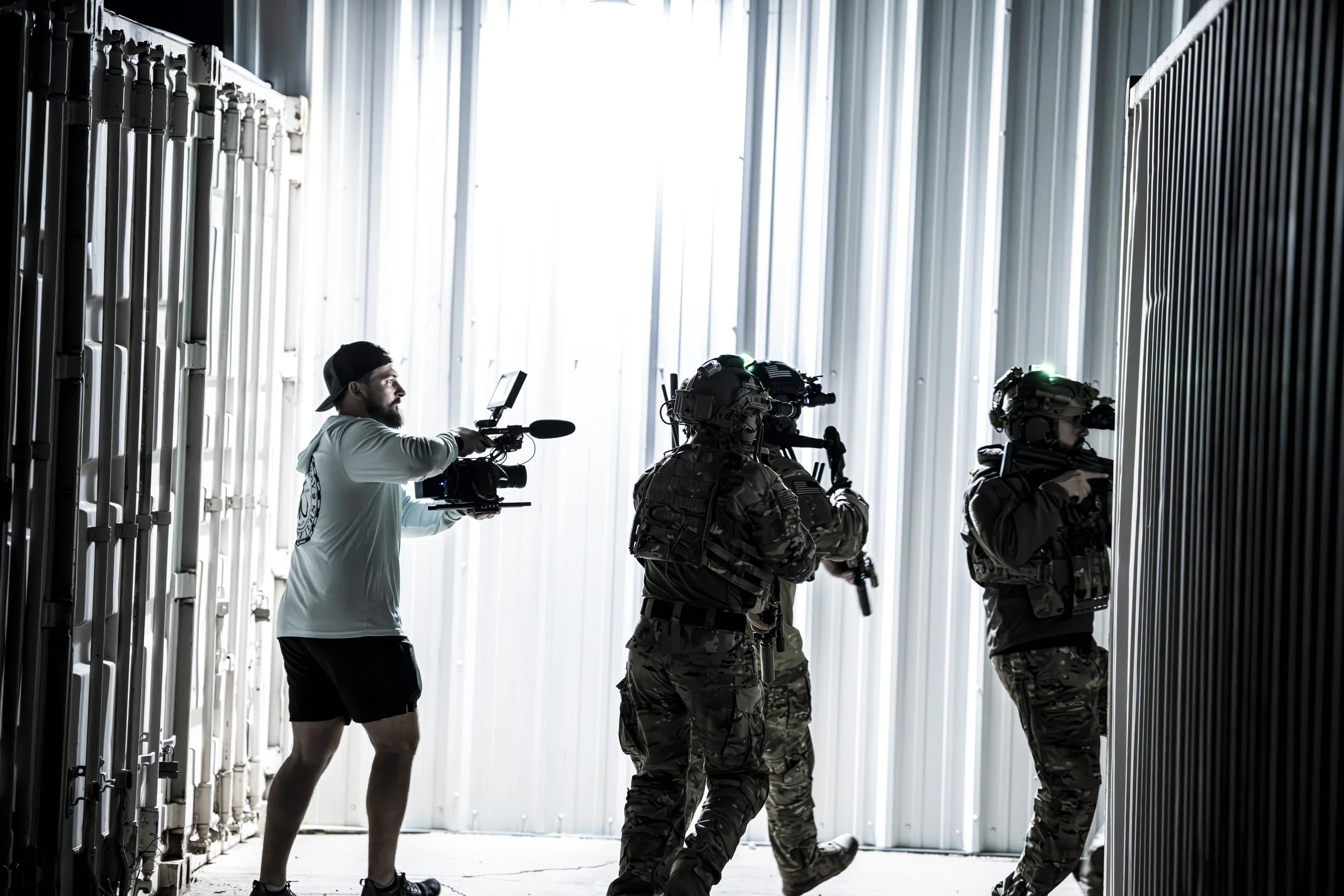 A film crew shooting a scene with armed soldiers in military gear inside a metal warehouse.